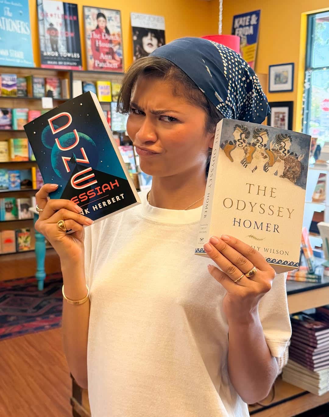A person in a blue headscarf and white shirt stands in a bookstore, holding up Dune Messiah and The Odyssey with a playful, quizzical look—like they're debating which story would get more stars on their favorite film app or Letterboxd.
