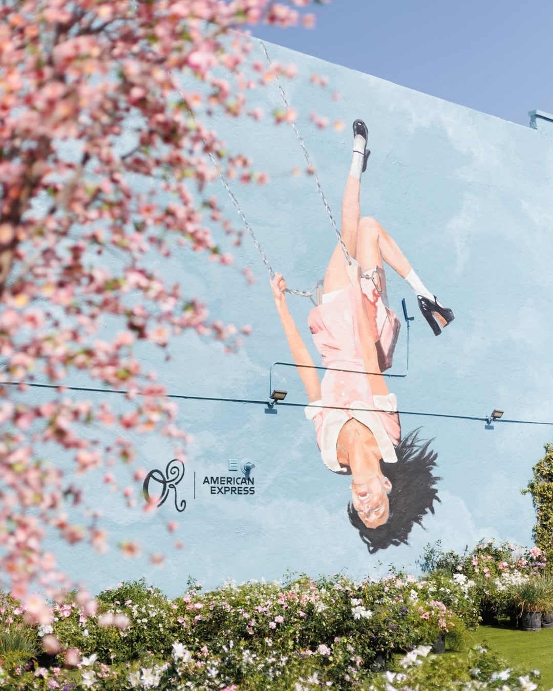 A colorful mural on a blue wall depicts a girl in a pink dress joyfully swinging upside down. Blossoming pink flowers and greenery appear in the foreground. The mural features the American Express logo.