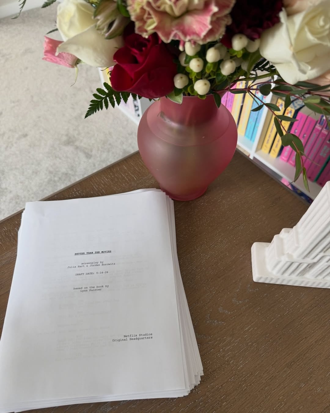 A stack of printed papers titled Beyond the Moon sits on a wooden desk beside a pink vase holding flowers. Colorful books, including trending BookTok YA adaptations, are visible on a shelf in the background.