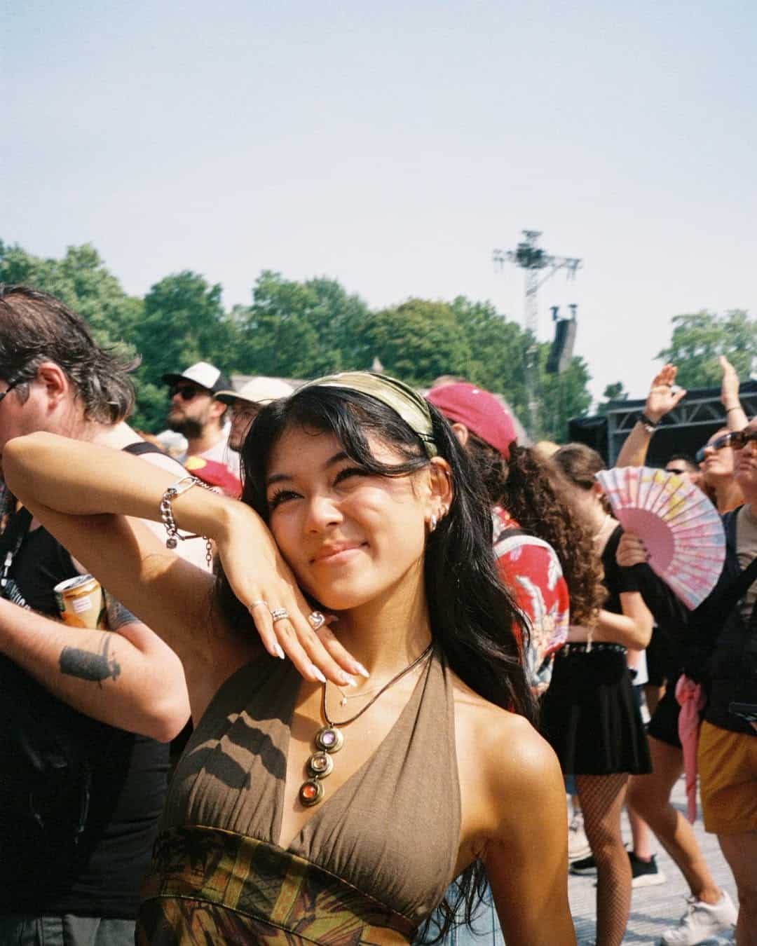 A smiling woman poses with one hand under her chin at an outdoor festival, wearing layered necklaces and a halter top—making the most of end-of-summer bucket list adventures. Around her, people enjoy drinks, a colorful fan, and sunny vibes near trees and stage equipment.