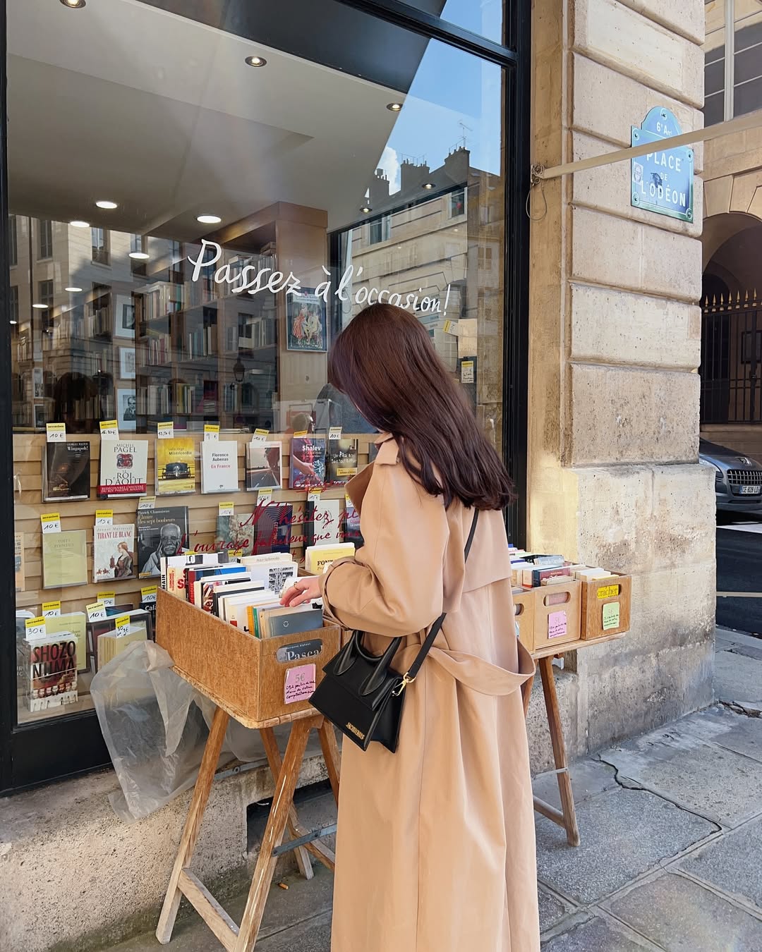 A woman in a long beige coat browses books displayed outside a bookstore on a city street, adding titles to her TBR as a stone building and the blue Place de l’Odéon sign appear in the background.