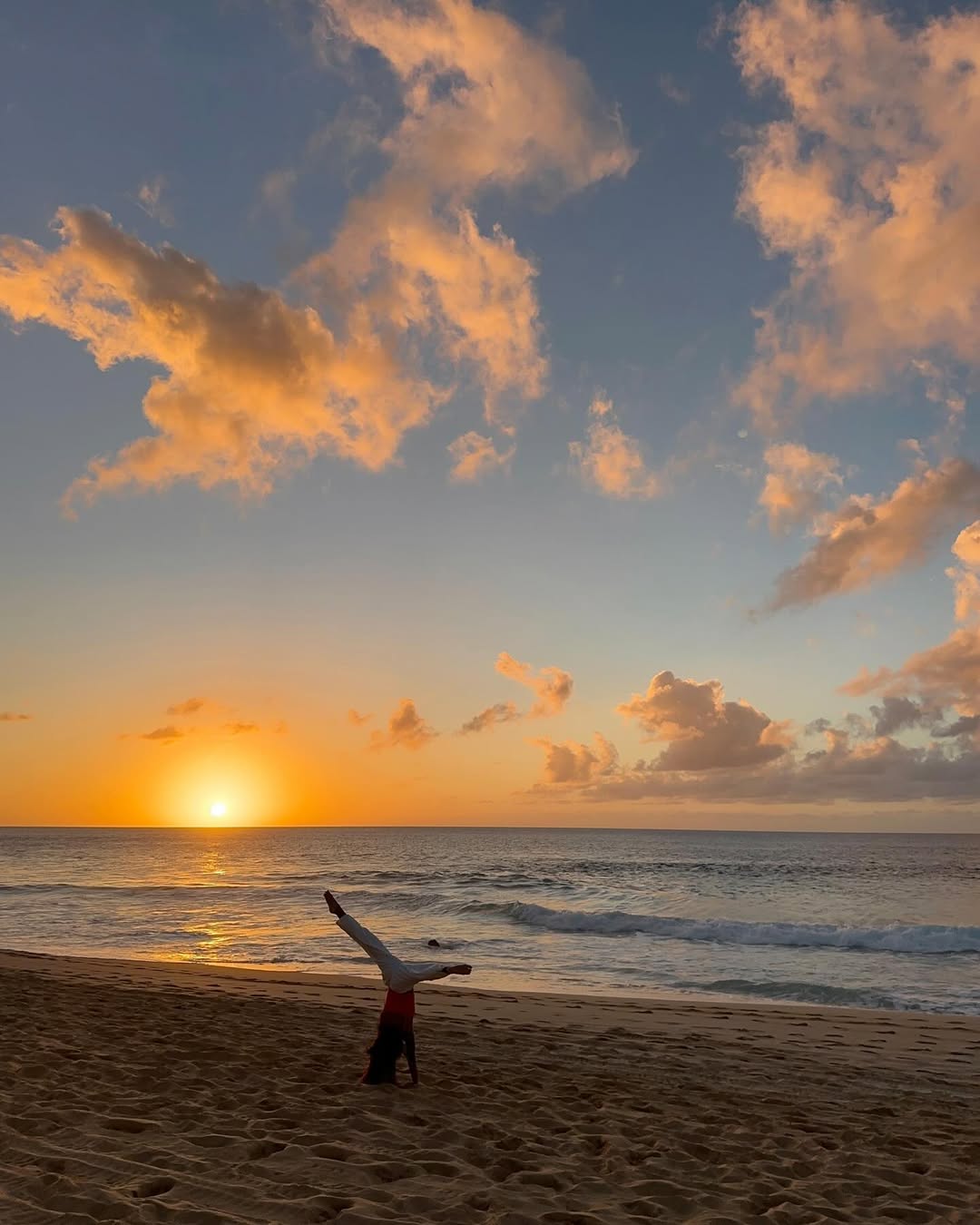 A person does a cartwheel on a sandy beach at sunset, with the sun low on the horizon and orange clouds scattered across the sky above the calm ocean—a perfect end-of-summer bucket list moment.