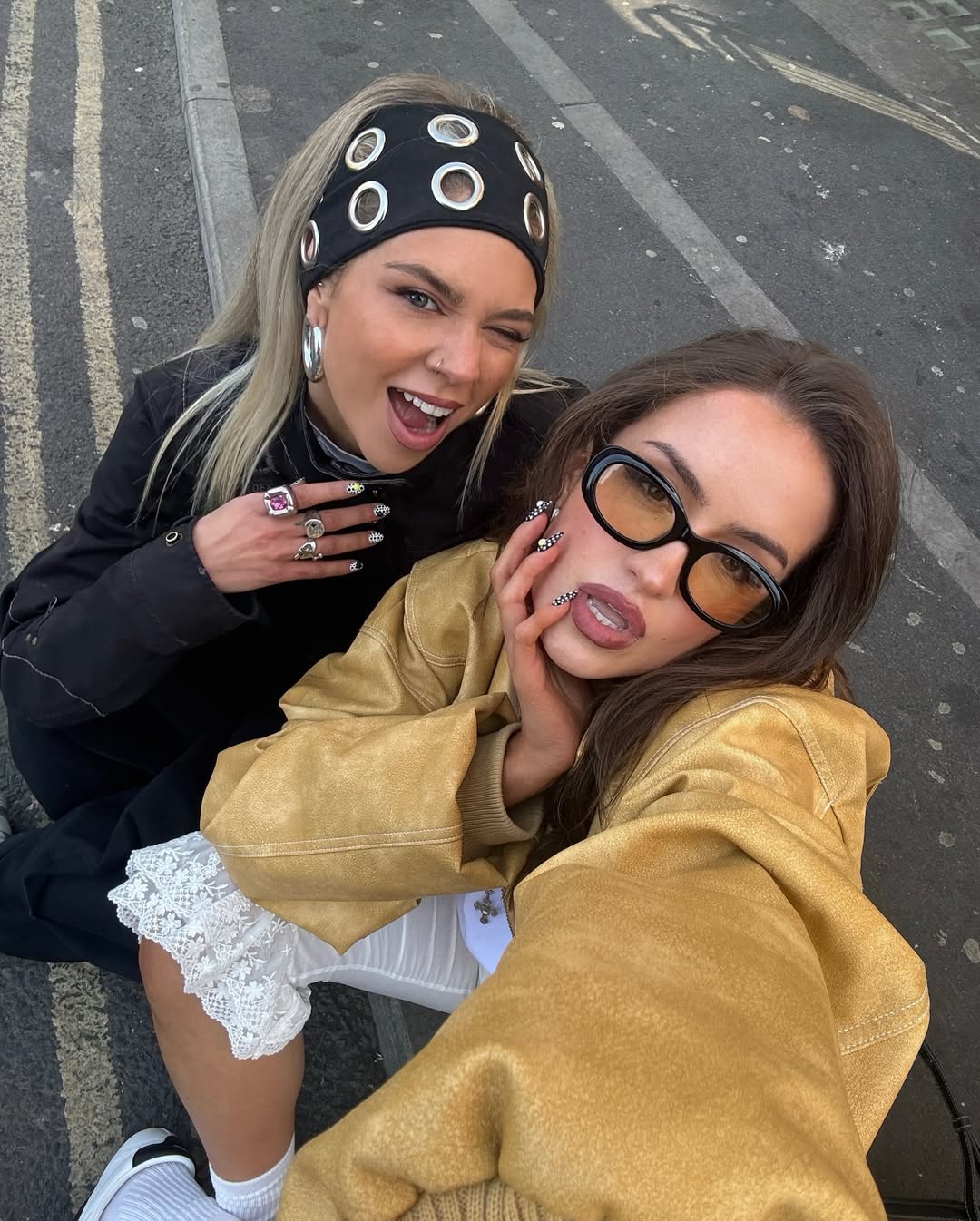 Two young women pose for a playful selfie on a street. One, sporting a stylish headband with large hoop earrings, grins beside her friend in sunglasses, a beige jacket, and white lace details. Both are making expressive faces.