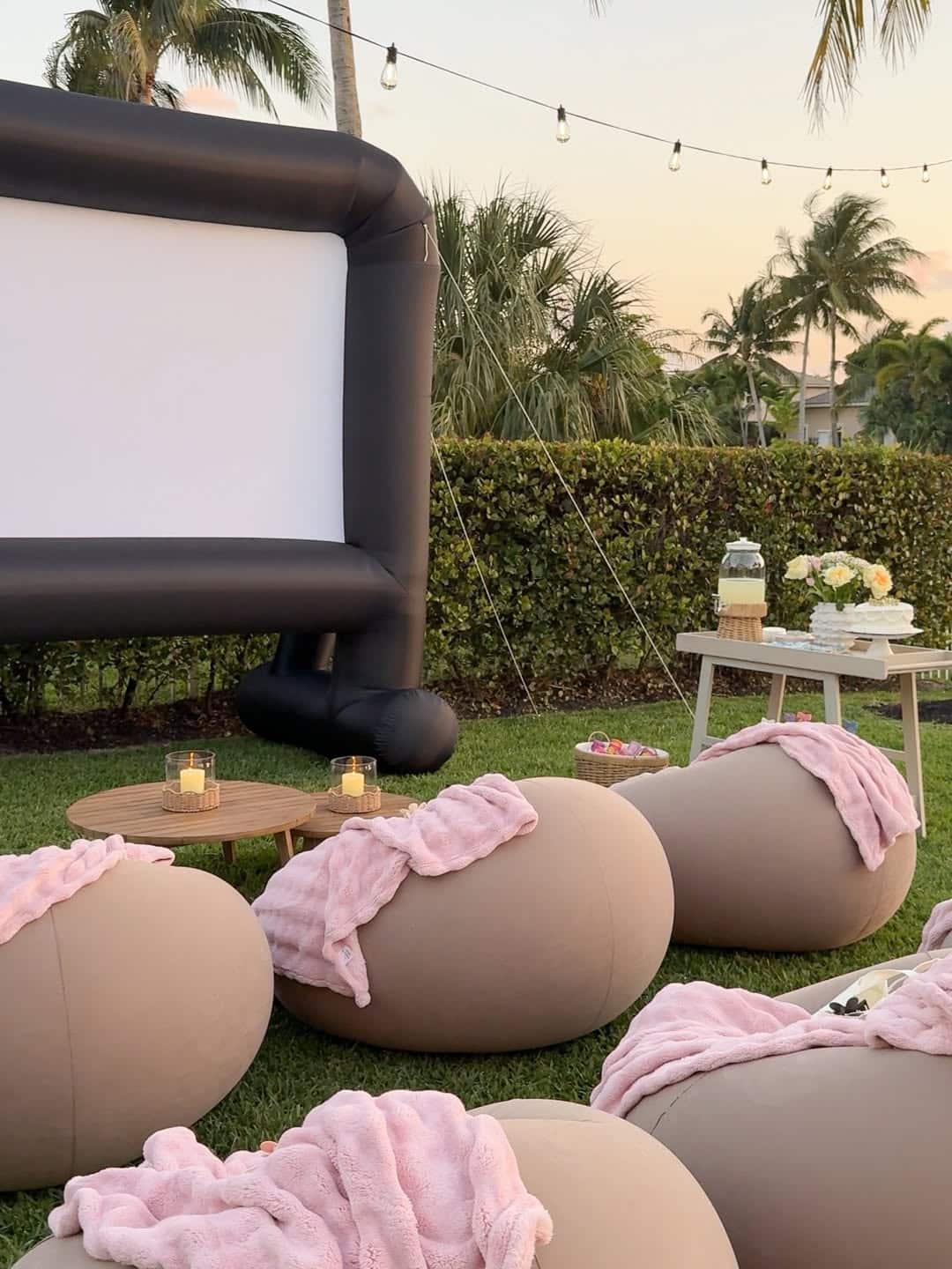 An outdoor movie setup perfect for your end-of-summer bucket list, featuring an inflatable screen, cozy beige seating cushions with pink blankets, low wooden tables with candles, a refreshment table, and string lights twinkling overhead on the grass.