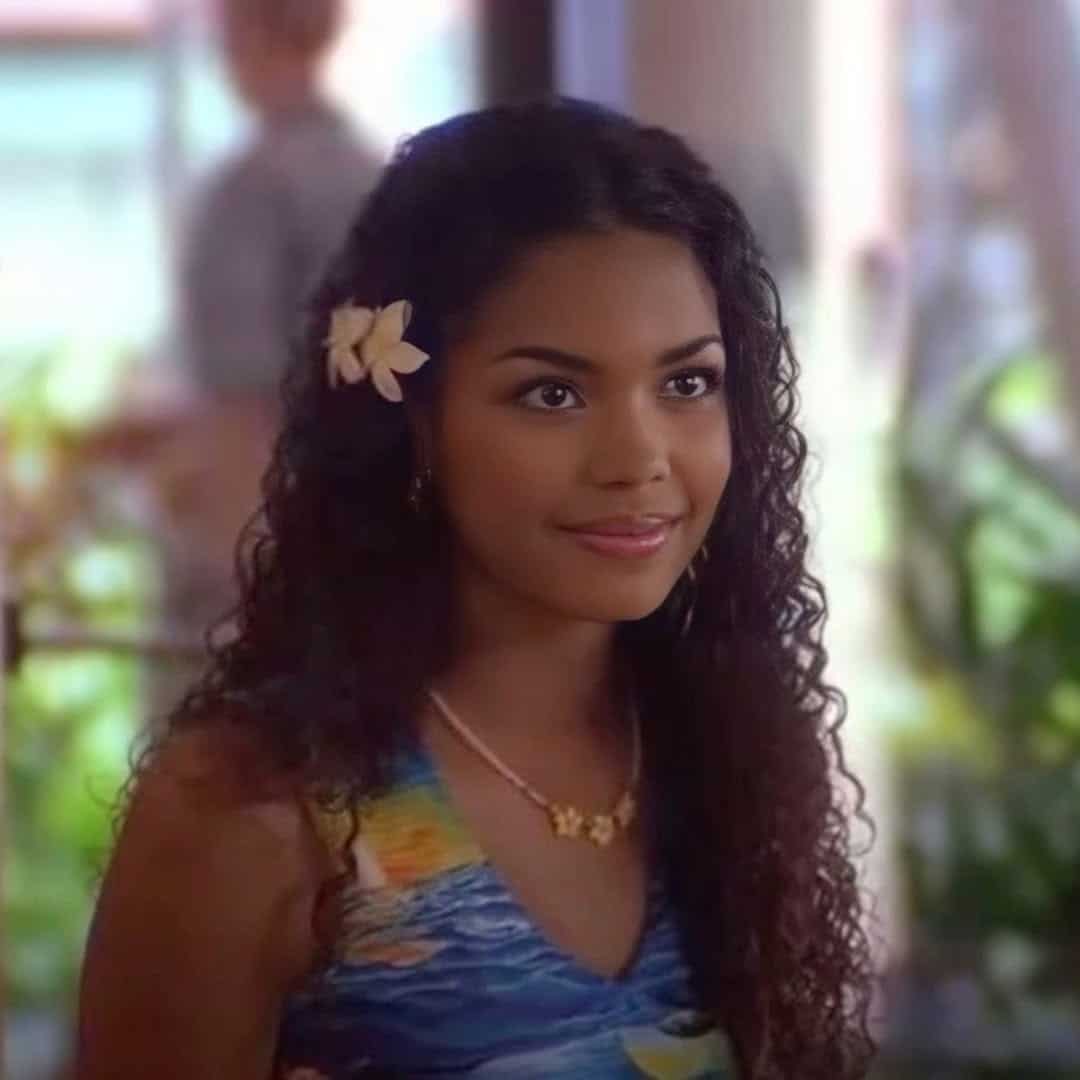 A young woman with long curly hair styled in a soft, celebrity hair look wears a flower behind her ear and a colorful dress, smiling indoors with blurred plants and people in the background.