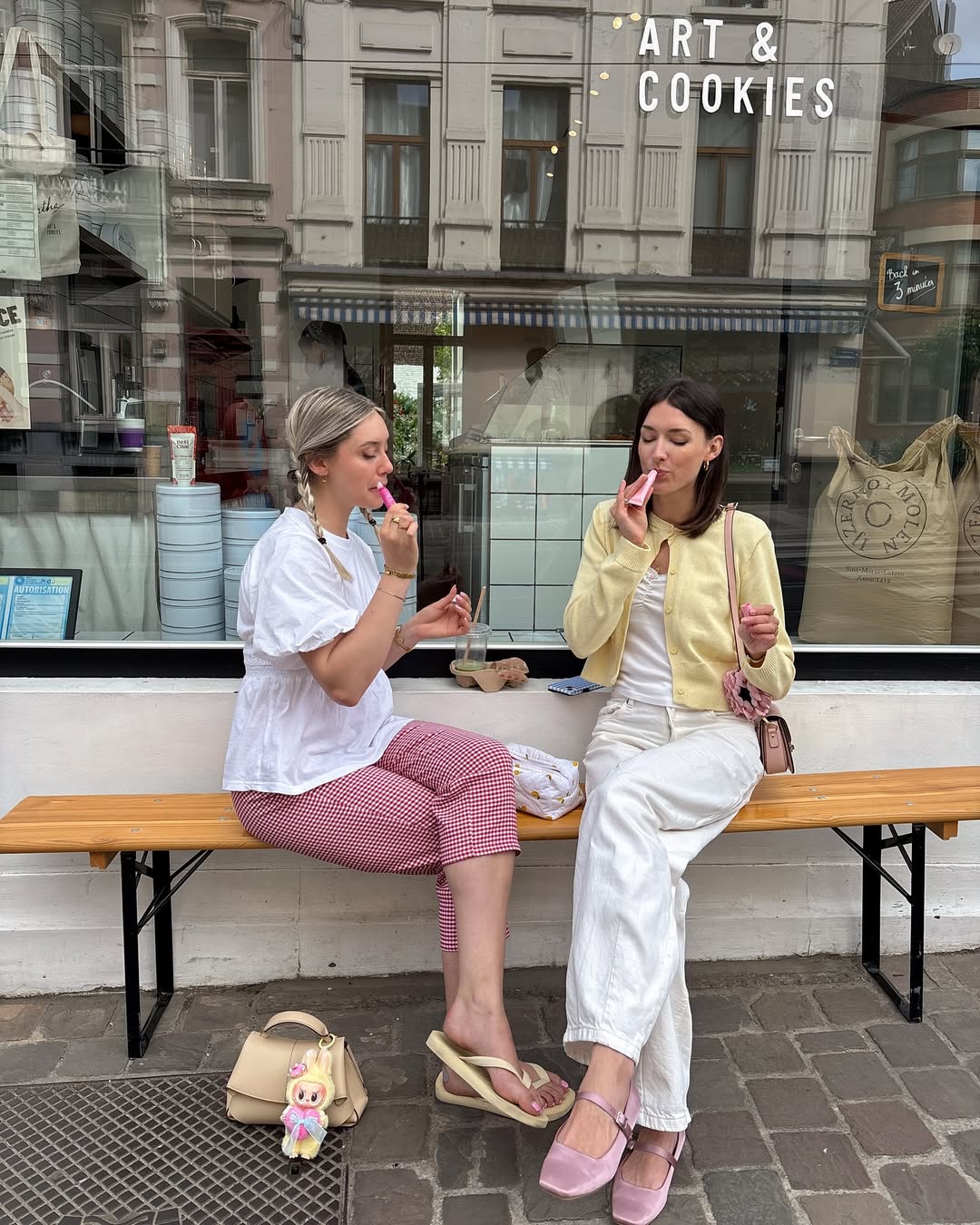 Two senior siblings sit on a wooden bench outside a bakery, enjoying pink popsicles. One wears a white blouse and red checkered pants, the other a yellow cardigan and white pants. Their purses rest nearby on the ground.