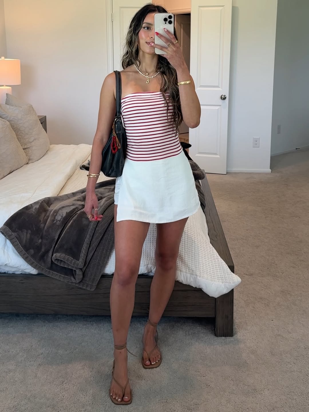 A woman stands in a bedroom taking a mirror selfie, showing off the ultimate Fourth of July outfit: a red and white striped tank top, white skirt, brown sandals, and black handbag. The room features a made bed, beige carpet, and neutral decor.