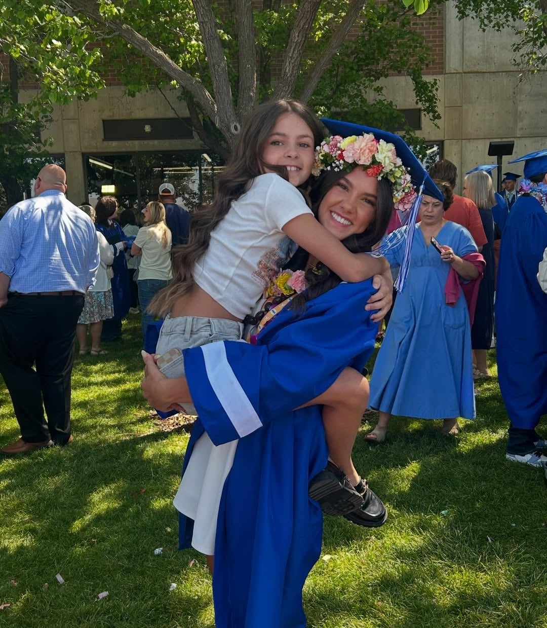 A smiling graduate in a blue cap and gown with a flower crown hugs and lifts her young sister at an outdoor graduation ceremony, celebrating the last summer with siblings, surrounded by others in similar gowns.