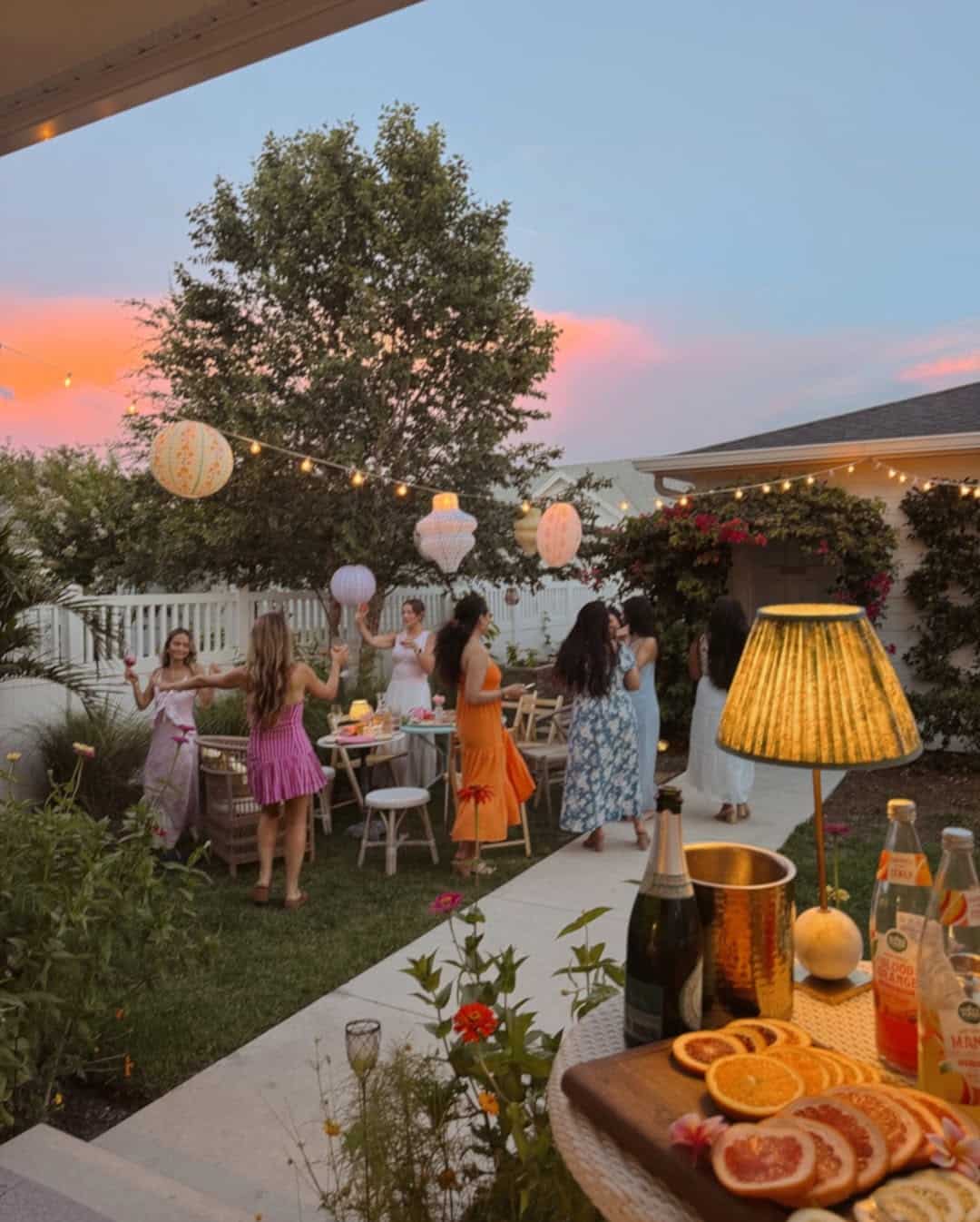 A group of women in colorful dresses gather in a backyard at sunset, surrounded by string lights and paper lanterns, reminiscing about last summer spent with senior siblings. Drinks, fruit, and a lamp sit on a table amid flowers and greenery.