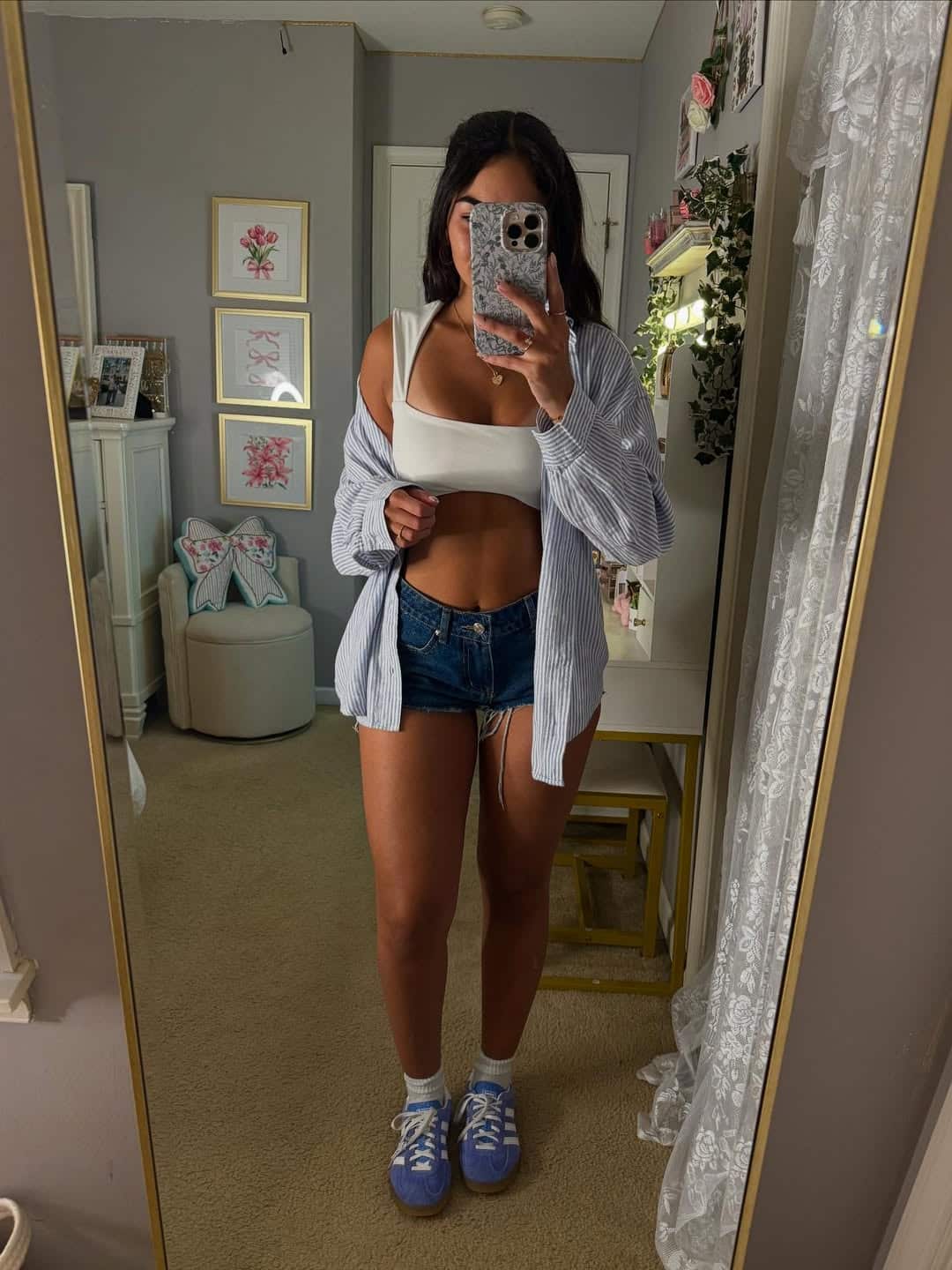 A woman takes a mirror selfie in a bedroom, wearing a white crop top, blue denim shorts, an open striped shirt, and purple sneakers—a casual Fourth of July outfit. The room features neutral decor, framed art, and a mirror with lace curtains.