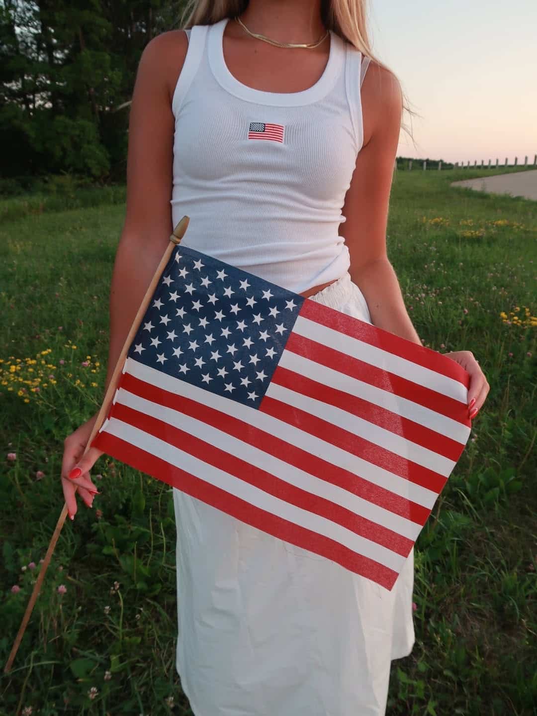 A woman stands outdoors on grass, wearing a white tank top and skirt—a perfect Fourth of July outfit—holding a small American flag. A tiny American flag is printed on her shirt, with wildflowers visible in the background.
