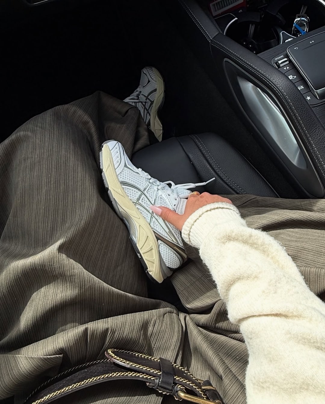 A person wearing a cream sweater, brown pleated skirt, and white sneakers sits in a car with legs crossed, resting a hand on their knee near a brown bag with a gold buckle—perfect inspiration for your Back-To-School shoes guide.
