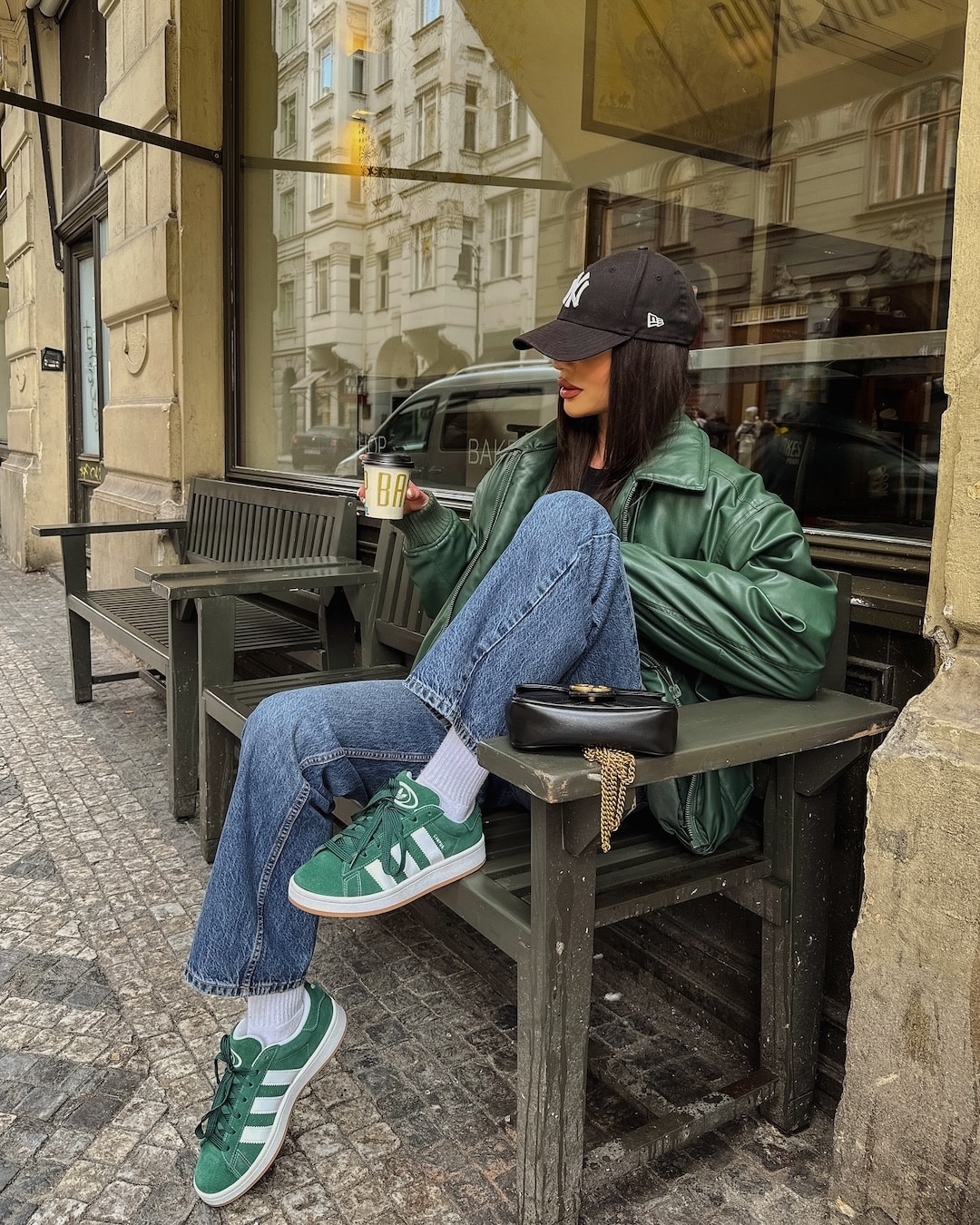 A woman in a green jacket, blue jeans, and green sneakers sits on a bench outside a café, holding a coffee cup. She wears a black cap and sunglasses, with a black handbag on the table—her look perfect for any Back-To-School Shoe Guide.