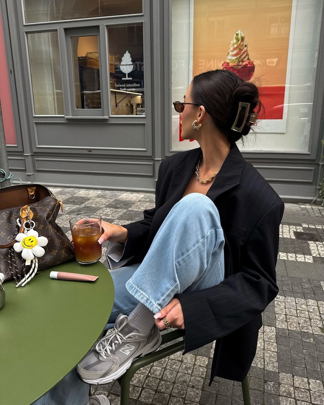 A woman in sunglasses and a black blazer sits at an outdoor café table with an iced drink, a designer bag, and a flower keychain. Her light blue jeans and gray sneakers make for effortless back-to-school style.