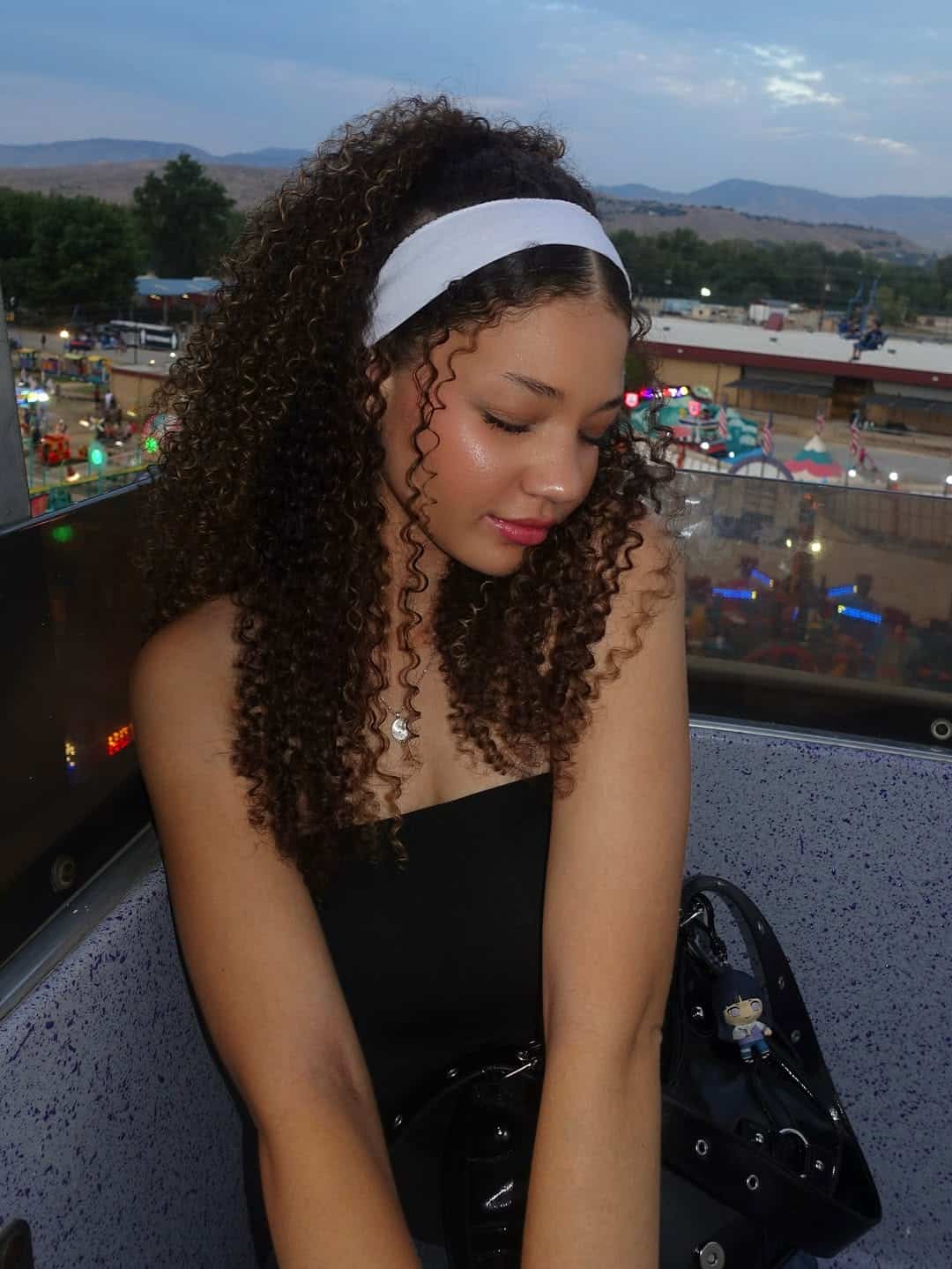 A young woman with curly hair and a white headband, one of her favorite hair accessories, sits in a Ferris wheel gondola at dusk, smiling with her eyes closed. A fairground with lights and rides glows in the background.