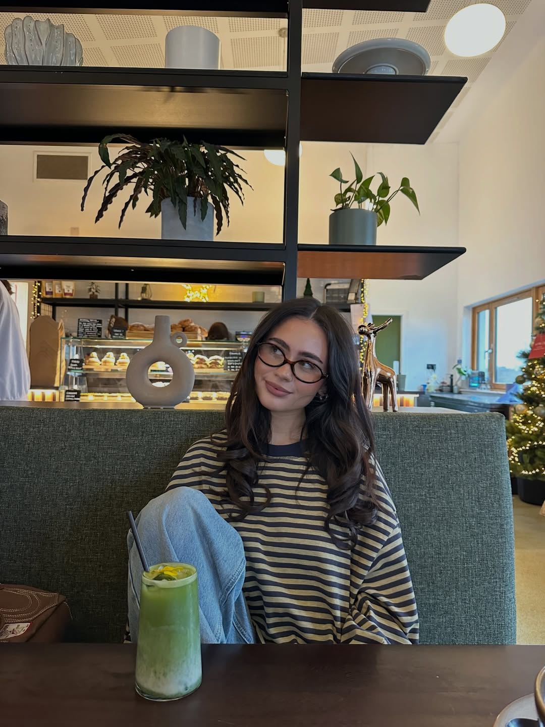 A woman with long brown hair and glasses, wearing a striped shirt and jeans, sits on a green bench in a cozy café. There is a green drink on the table and plants and shelves in the background.