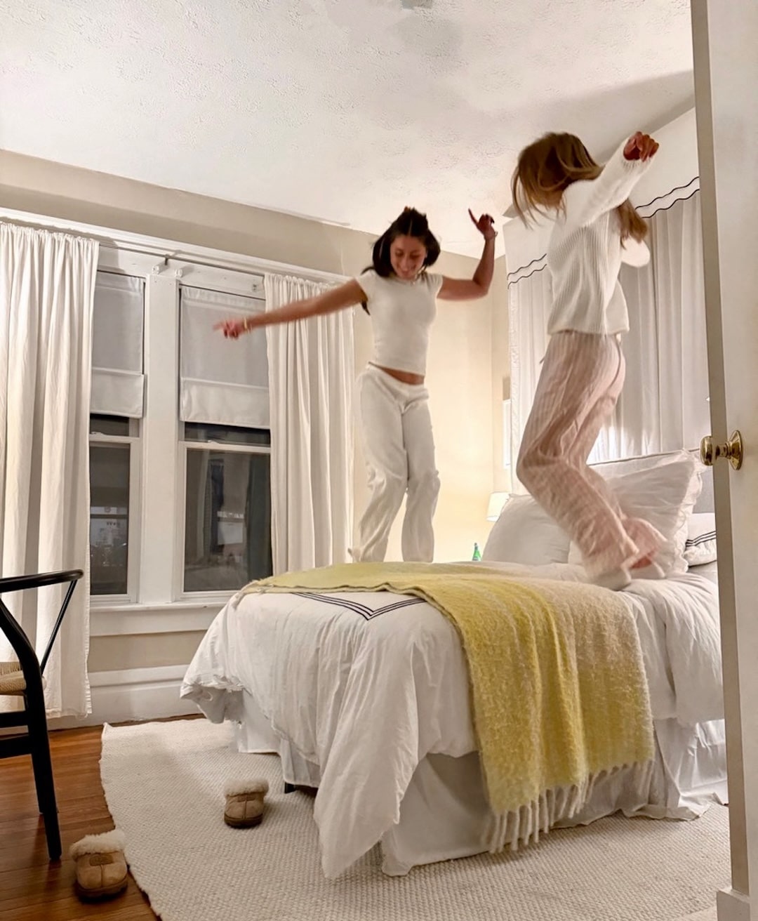 Two women in pajamas are joyfully jumping on a bed in a cozy, well-lit bedroom, reliving the fun of spending summer with siblings. A yellow blanket is draped across the bed and slippers rest on the floor.