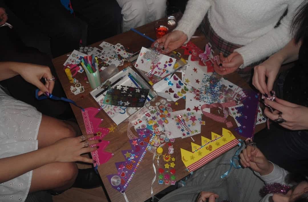 Several people, including senior siblings, sit around a table crafting colorful paper crowns decorated with stickers, rhinestones, and markers. Various art supplies and decorations are scattered across the tabletop.
