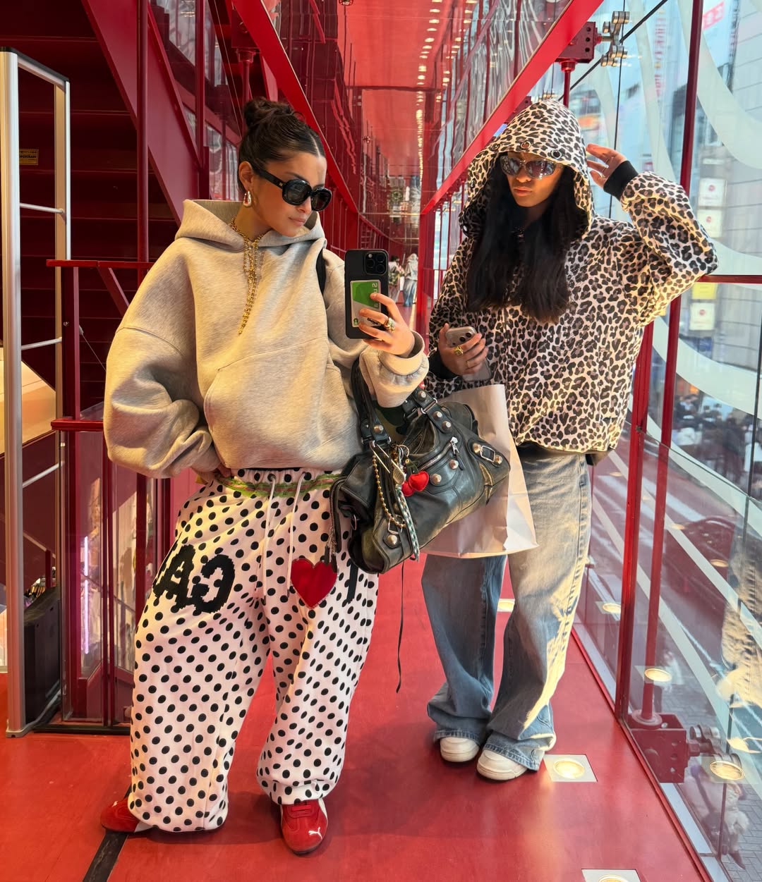 Two women pose in a modern, red glass hallway. One wears a gray hoodie, polka dot pants, and red shoes; the other wears a leopard hoodie, baggy jeans, and white sneakers. Both wear sunglasses and hold phones.