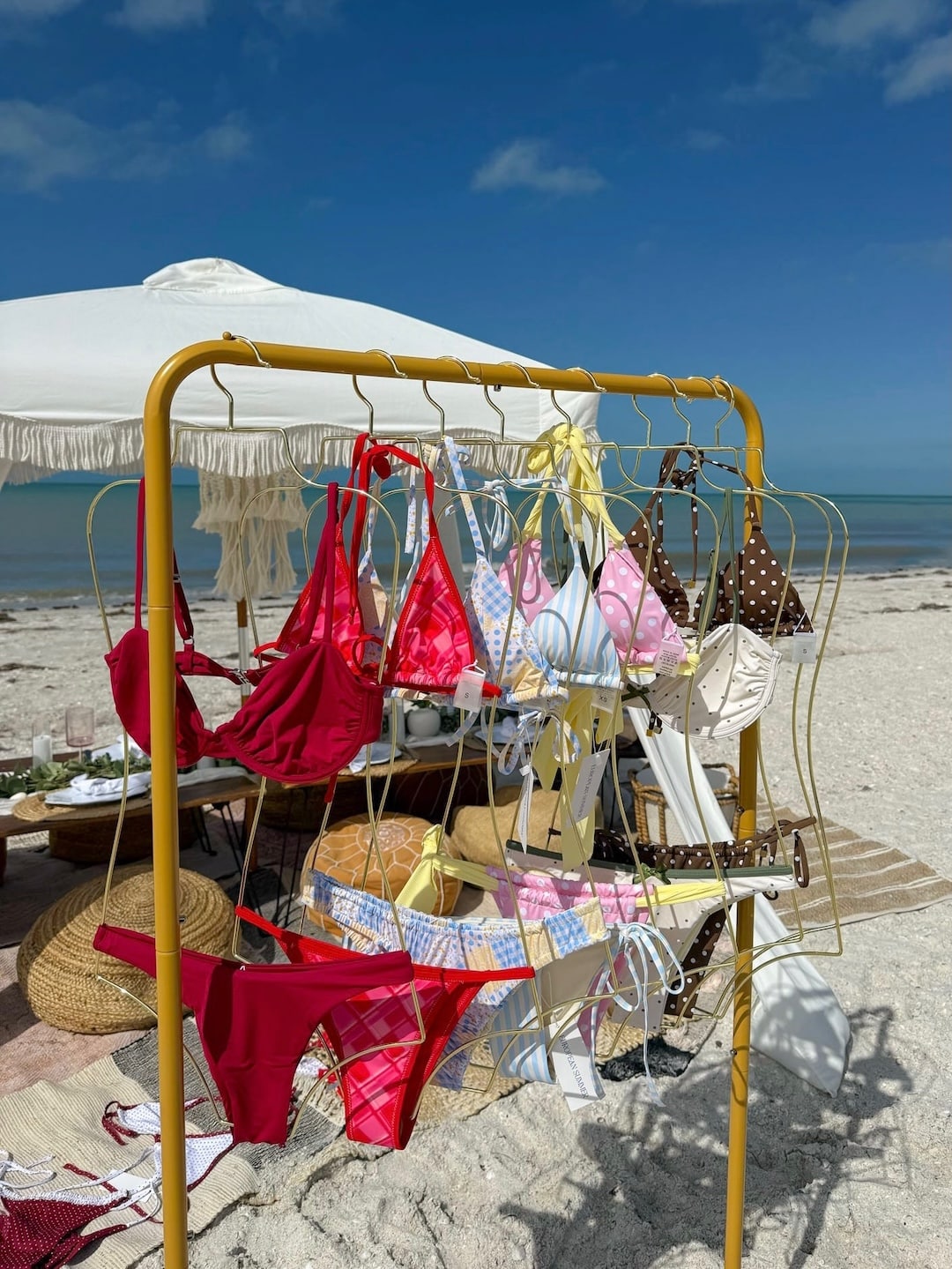 A gold rack displays colorful bikinis in red, pink, white, yellow, and brown polka dots on a sandy beach. In the background, there’s a white parasol, picnic setup, and the calm sea under a blue sky.