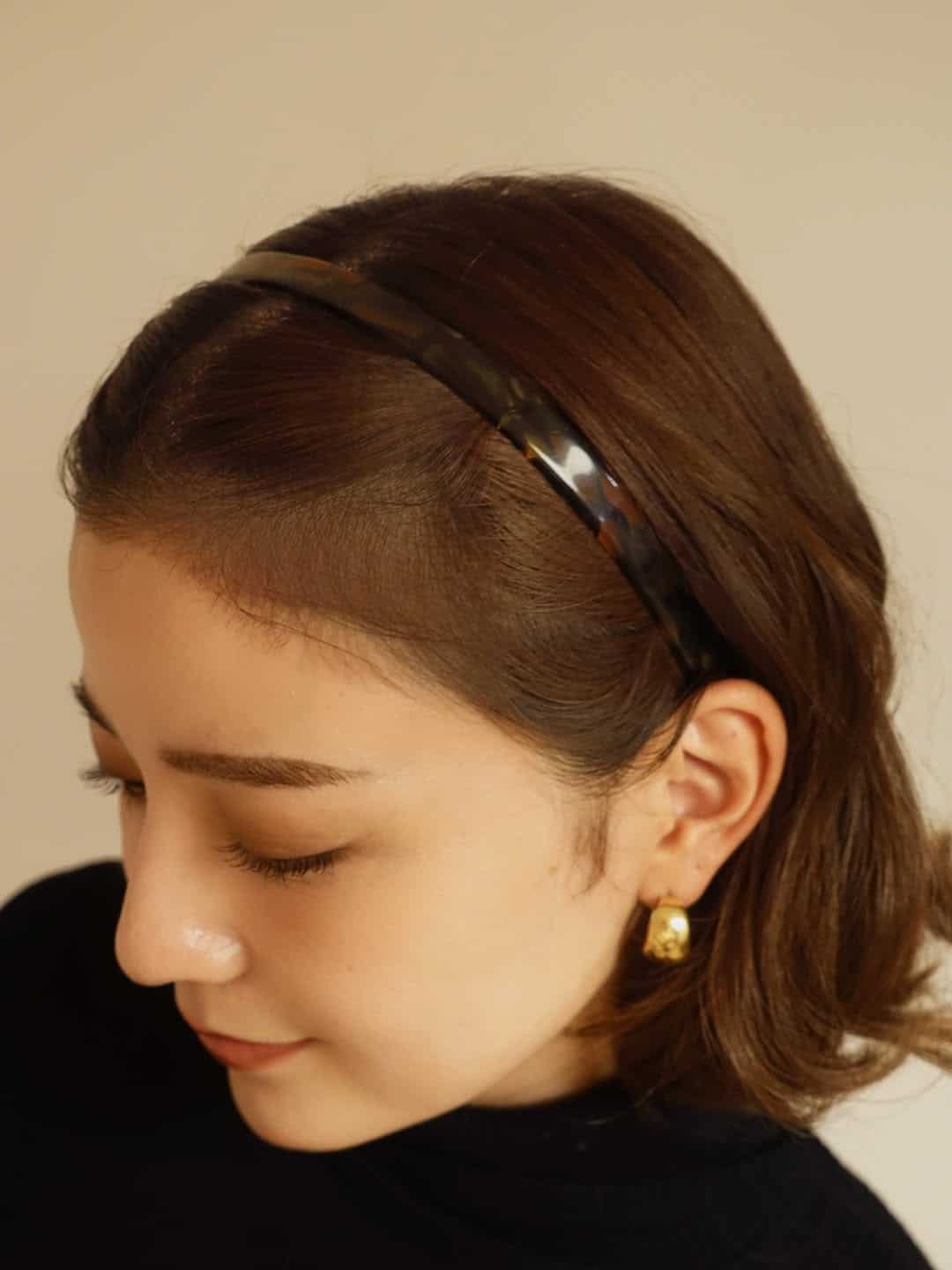A woman with short brown hair wears a black headband and gold earrings. Captured in this headband edit, she looks down and smiles softly, dressed in a black top against a neutral background.