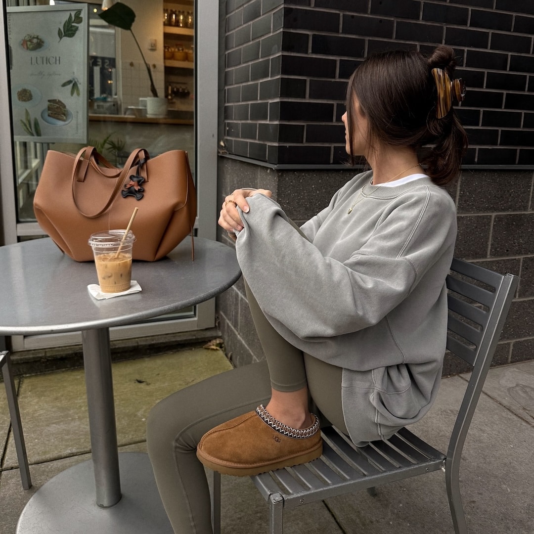 A woman in an oversized grey sweatshirt, green leggings, and brown slippers sits on a metal chair at an outdoor café table with an iced coffee and a large tan handbag—a cozy look perfect for any shoe guide or finding your ideal back to school shoes.