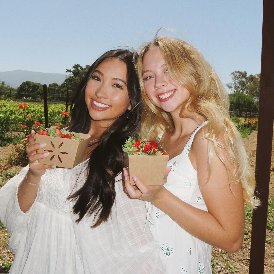 Two young women, possibly senior siblings, smiling and holding small boxes of strawberries, standing outdoors in a sunny garden or farm with flowers and greenery—a perfect snapshot of summer activities.