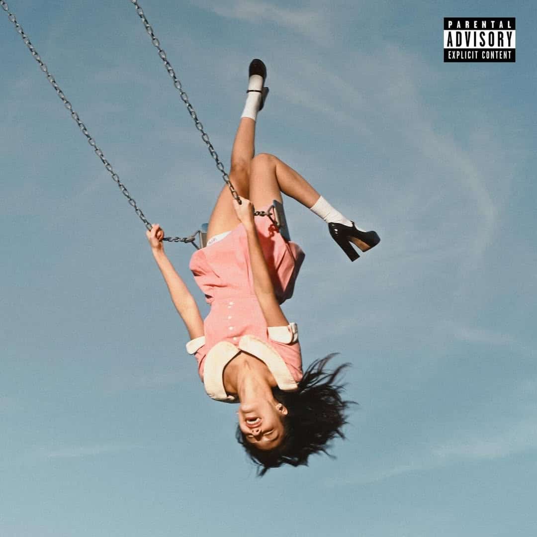 A young woman in a pink dress swings upside down against a blue sky, her hair hanging down and legs in the air. The Parental Advisory: Explicit Content label appears as her zodiac sign and "You Seem Pretty Sad for a Girl So in Love" stream through the scene.