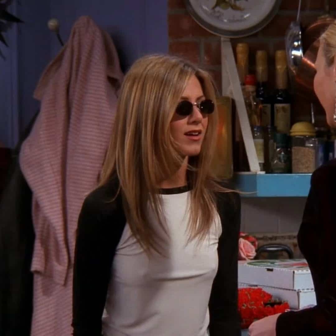 A woman with long straight hair, showcasing modern hairstyles, wears black sunglasses and a white and black raglan shirt. She stands indoors talking to another person, with kitchen items and a pink striped robe visible in the background.