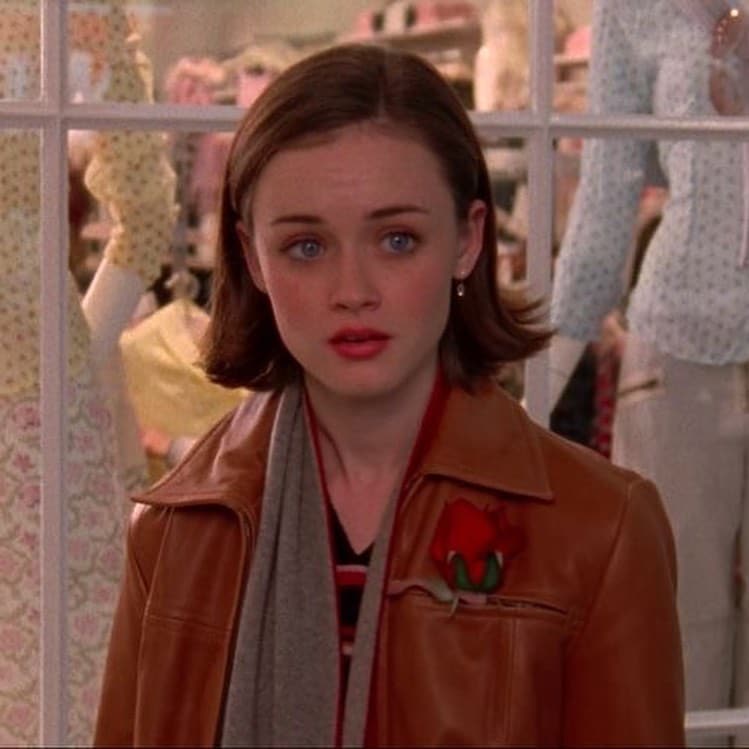 A young woman with short brown hair styled in a trendy salon hairstyle, wearing a brown leather jacket and a rose pin, stands indoors in front of a window display with mannequins dressed in patterned clothes. She looks surprised or concerned.