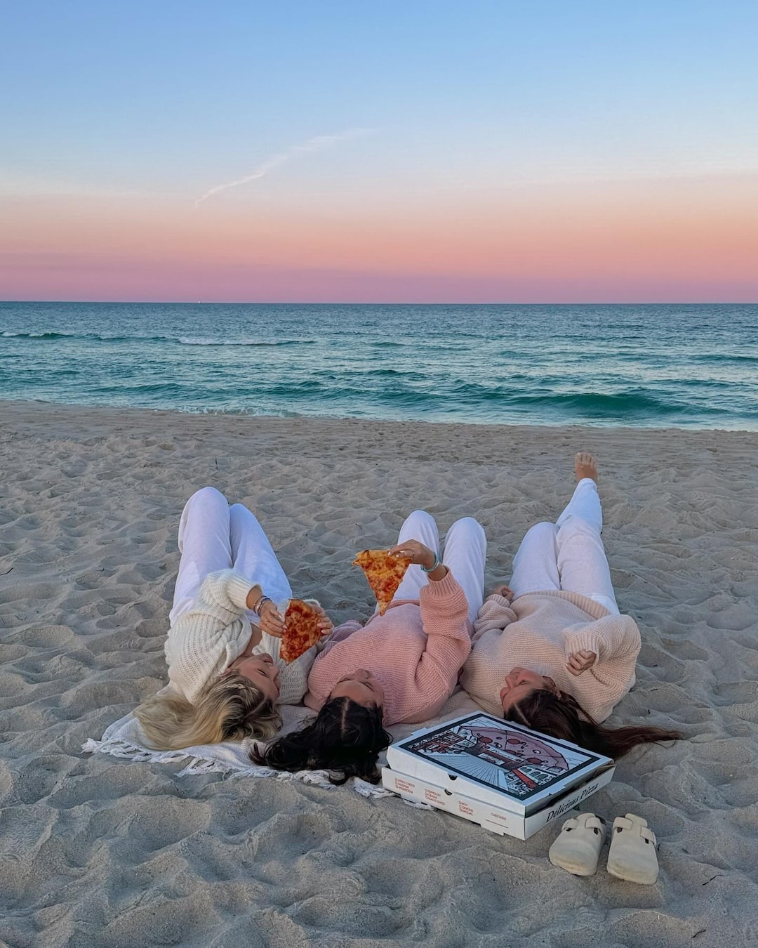 Three people in cozy loungewear lie on the sand at the beach, eating pizza from an open box at sunset. The sky is pink and blue over the calm ocean in the background.