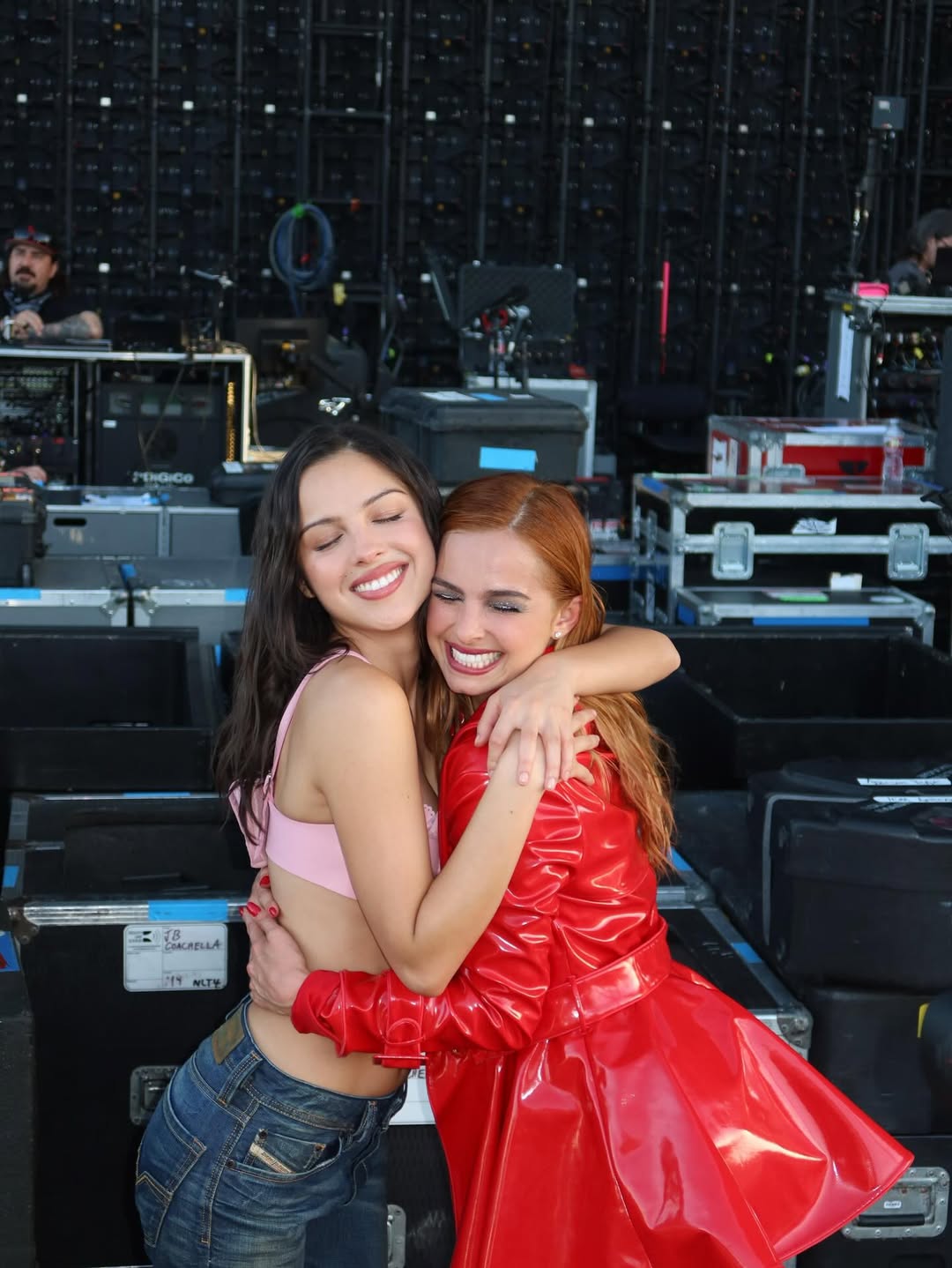 Two women are hugging and smiling with their eyes closed. One wears a pink top and jeans; the other sports a bright red coat. They stand backstage with equipment and cables—maybe sharing favorite love songs before a music streaming show.