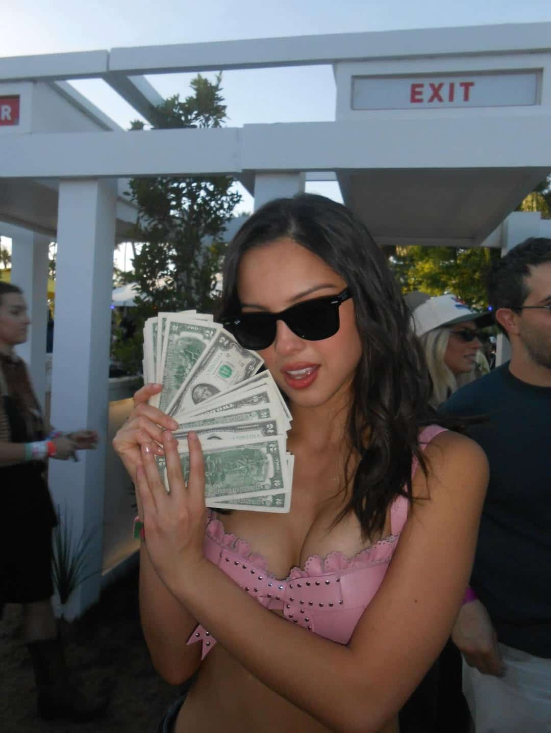A woman wearing black sunglasses and a pink top holds a fan of U.S. dollar bills in both hands, standing outdoors near EXIT signs—her confidence shining brighter than her zodiac sign as if she’s streaming "You Seem Pretty Sad for a Girl So in Love.