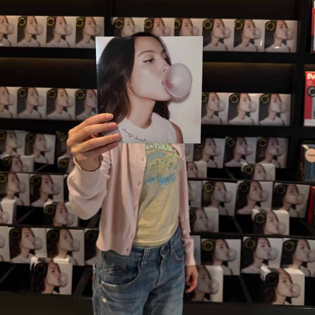 A person holds an album cover in front of their face, aligning the image of a woman blowing bubblegum with their own head. Behind them, stacked albums echo the same cover—perfect for anyone in love with music and looking to stream the latest track.