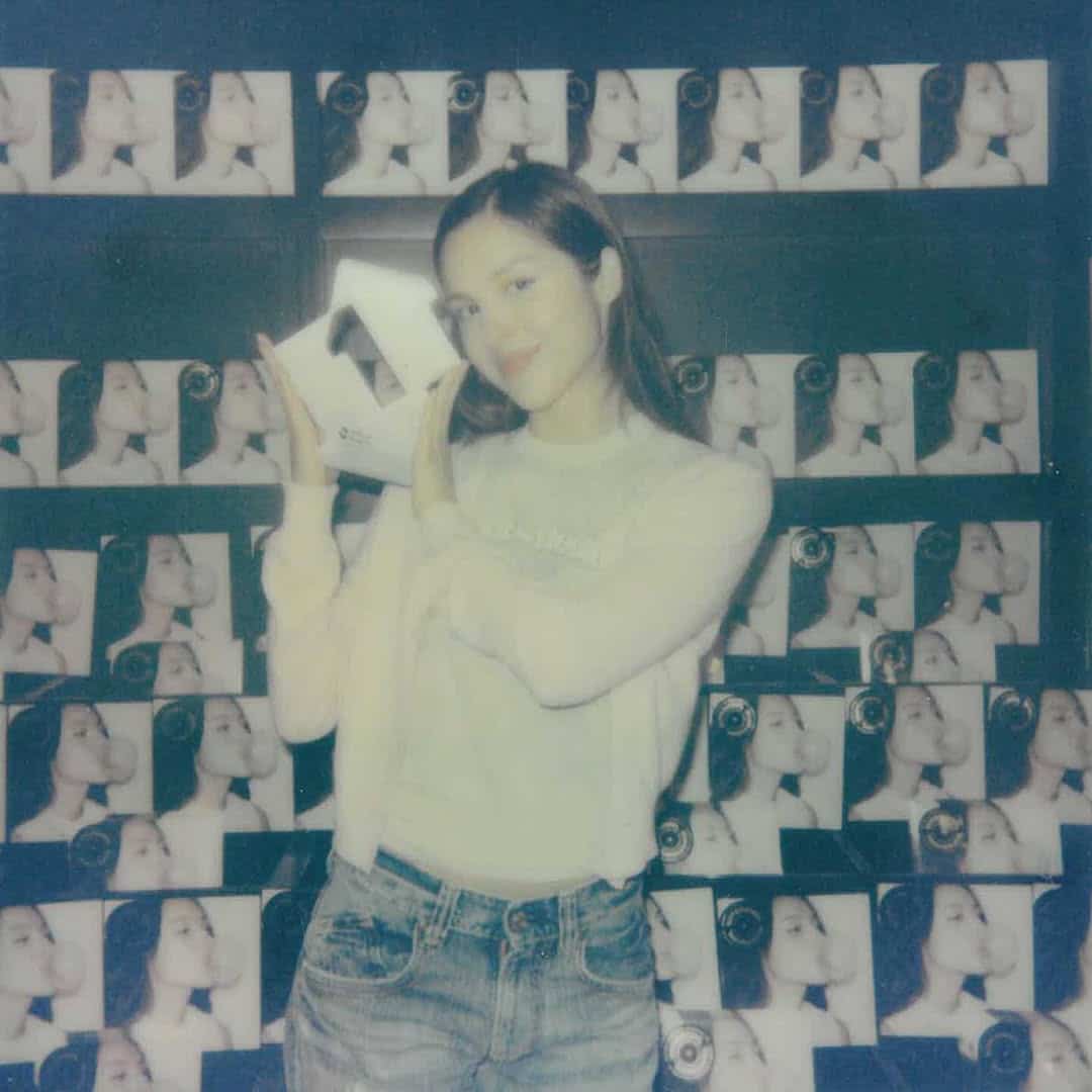 A young woman smiles and holds up a product box in front of a wall covered with repeated images of a woman's side profile, her zodiac sign delicately visible on her necklace. She wears a light top and jeans in a softly lit, slightly blurry photo.