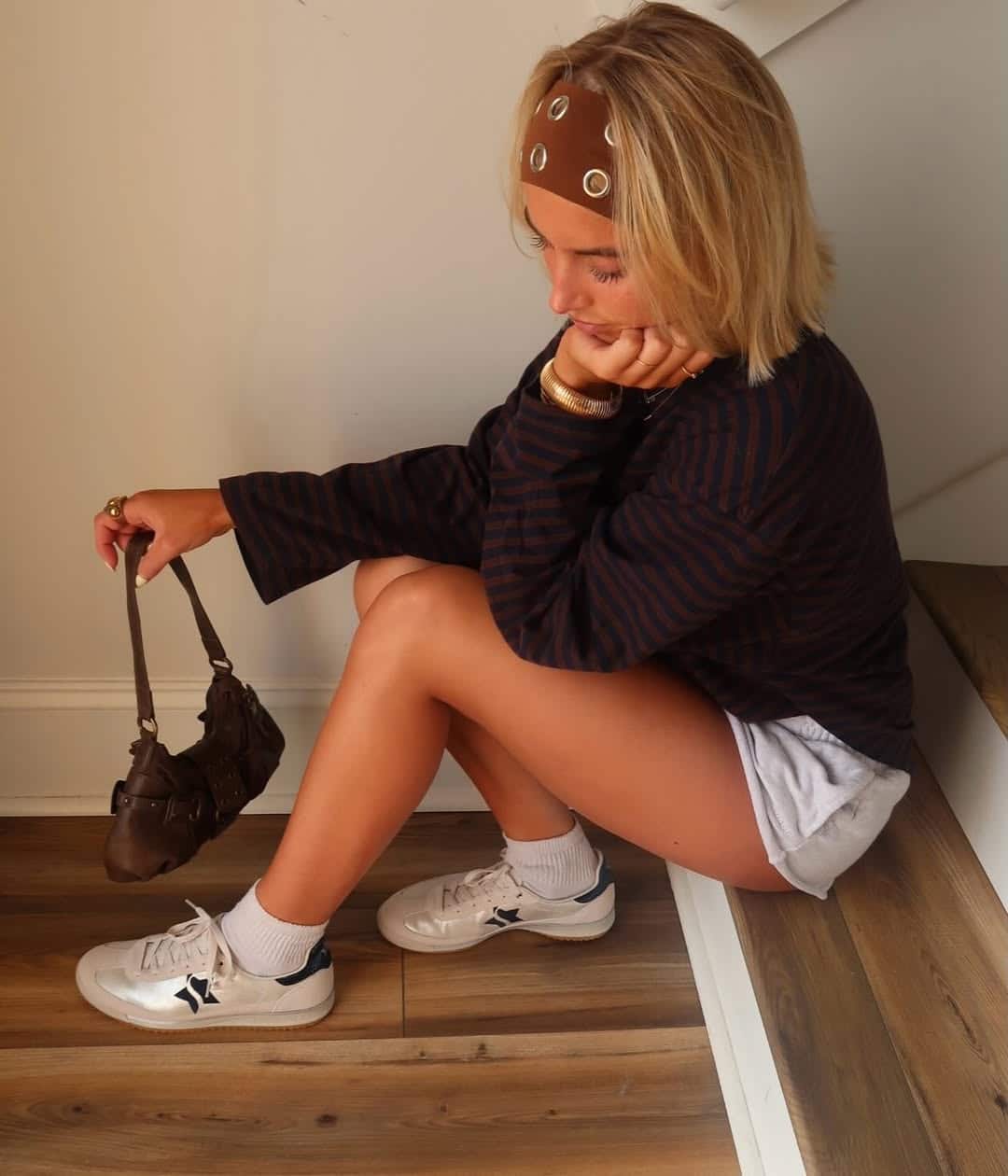 A person with blonde hair sits on wooden stairs, resting their chin on their hand. They wear a brown headband, a striped long-sleeve top, white shorts, white sneakers, and hold a small brown handbag—a chic headband edit.