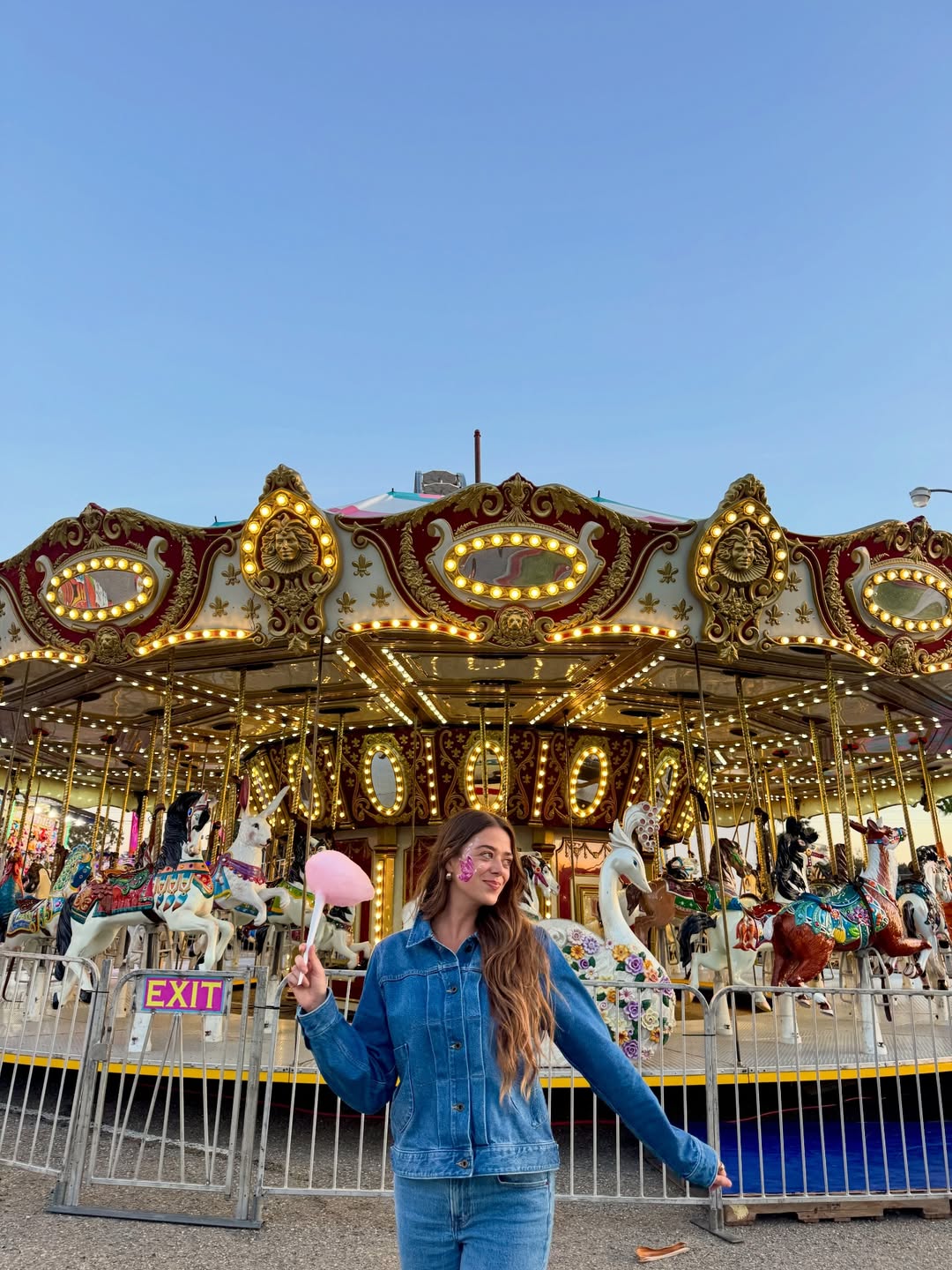 A young woman in a denim jacket holds pink cotton candy and smiles in front of a brightly lit, ornate carousel—a perfect bucket list moment for anyone seeking classic summer activities under a clear blue sky.