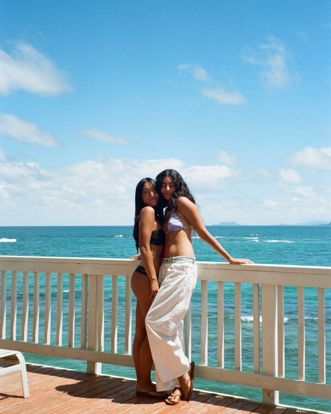 Two women in swimwear stand close together on a wooden deck by the ocean, leaning against a white railing. With blue sky and clouds overhead, they enjoy scenic views—soaking up an end-of-summer moment on their bucket list of summer activities.