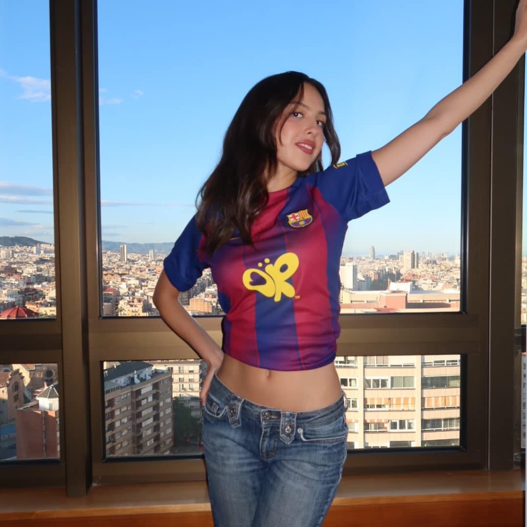 A woman wearing a Barcelona soccer jersey and jeans poses with one arm raised in front of a large window, tracking the cityscape and blue sky behind her.