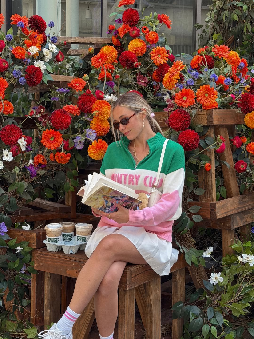 A woman wearing sunglasses, a green and pink Country Club sweater, and a white skirt sits on a wooden bench, reading a book from her TBR. Behind her are vibrant orange, red, and purple flowers. Four coffee cups sit on the bench.