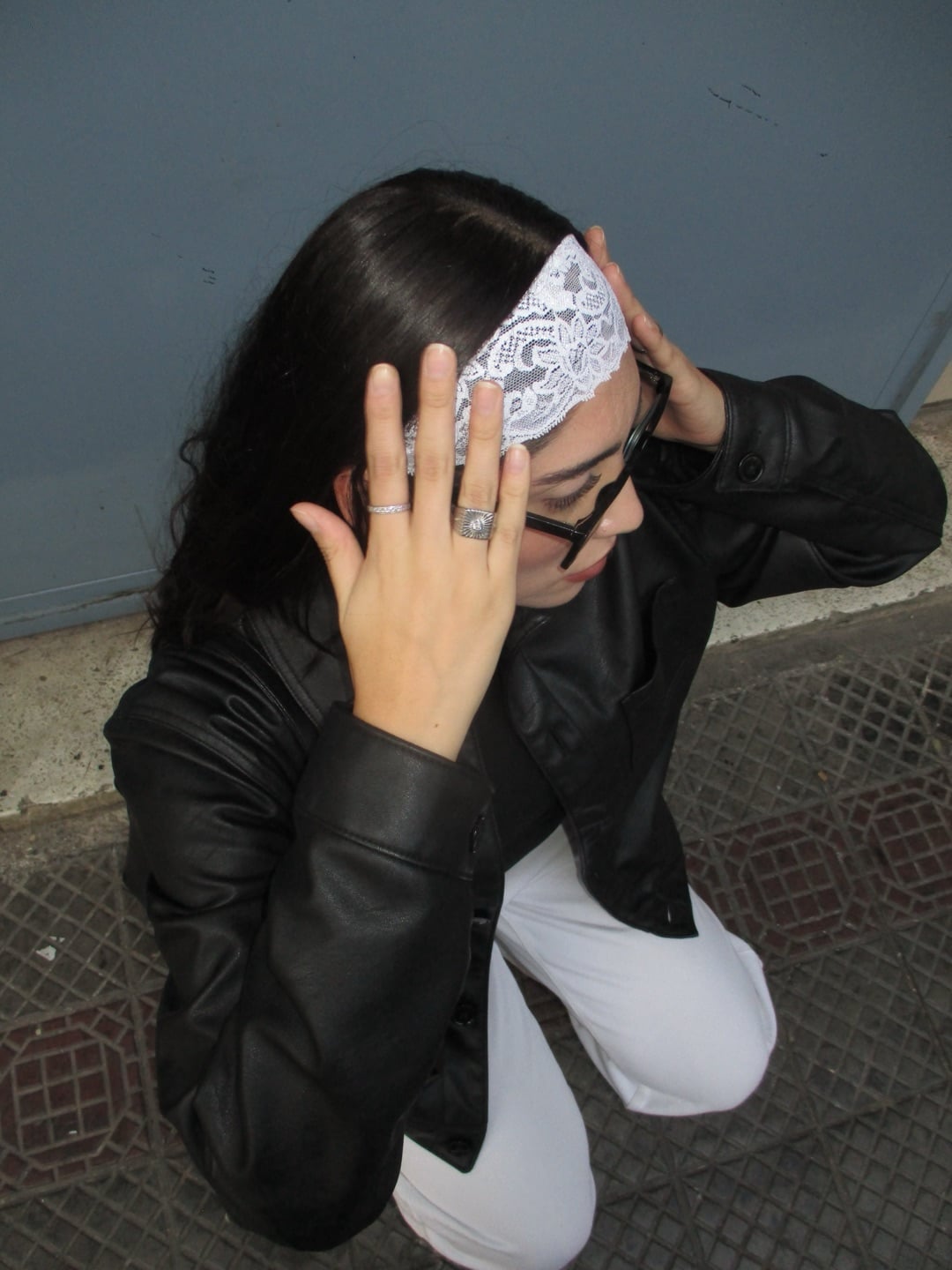 A person wearing a black jacket, white pants, and a white lace headband edit kneels on a patterned sidewalk, holding their head with both hands. Their hair is dark and they wear stylish sunglasses.