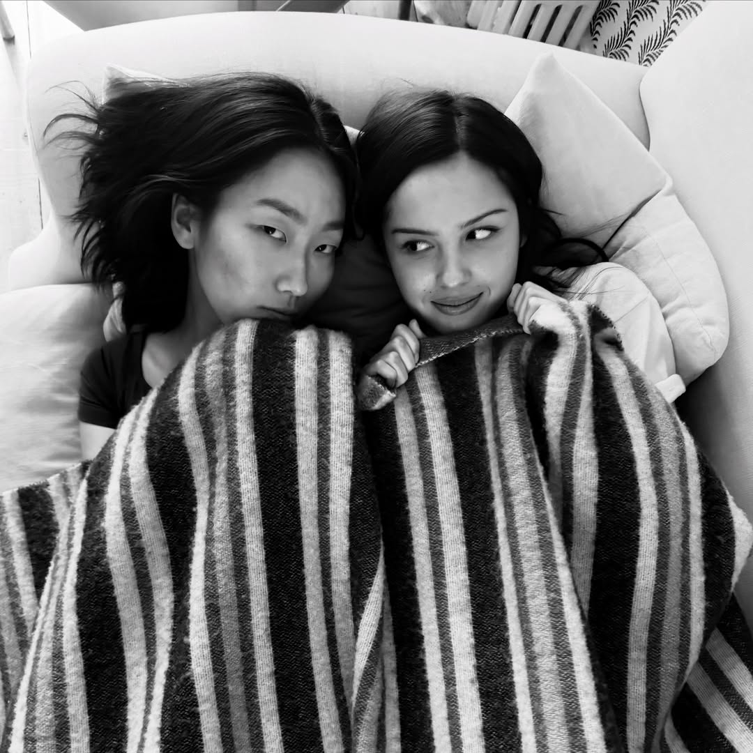 Two young women lie close together on a couch, partially covered by a striped blanket, looking at each other playfully as a love song streams softly in the background. The photo is in black and white.