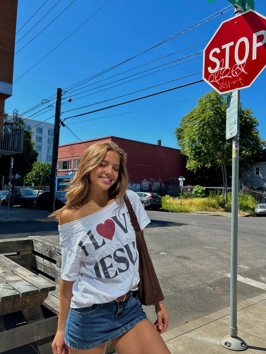 A woman with light brown hair smiles outdoors on a sunny day, wearing a white off-shoulder I ♥ JESUS t-shirt and denim shorts, standing near a stop sign at a street corner—embracing vacation at home vibes.