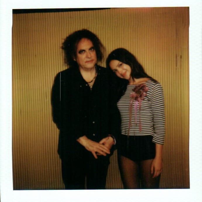 A man dressed in black with messy hair stands next to a smiling young woman in a striped shirt and shorts, who leans her head on his shoulder. Their zodiac signs might match as perfectly as they do, posed together before a yellowish wall.