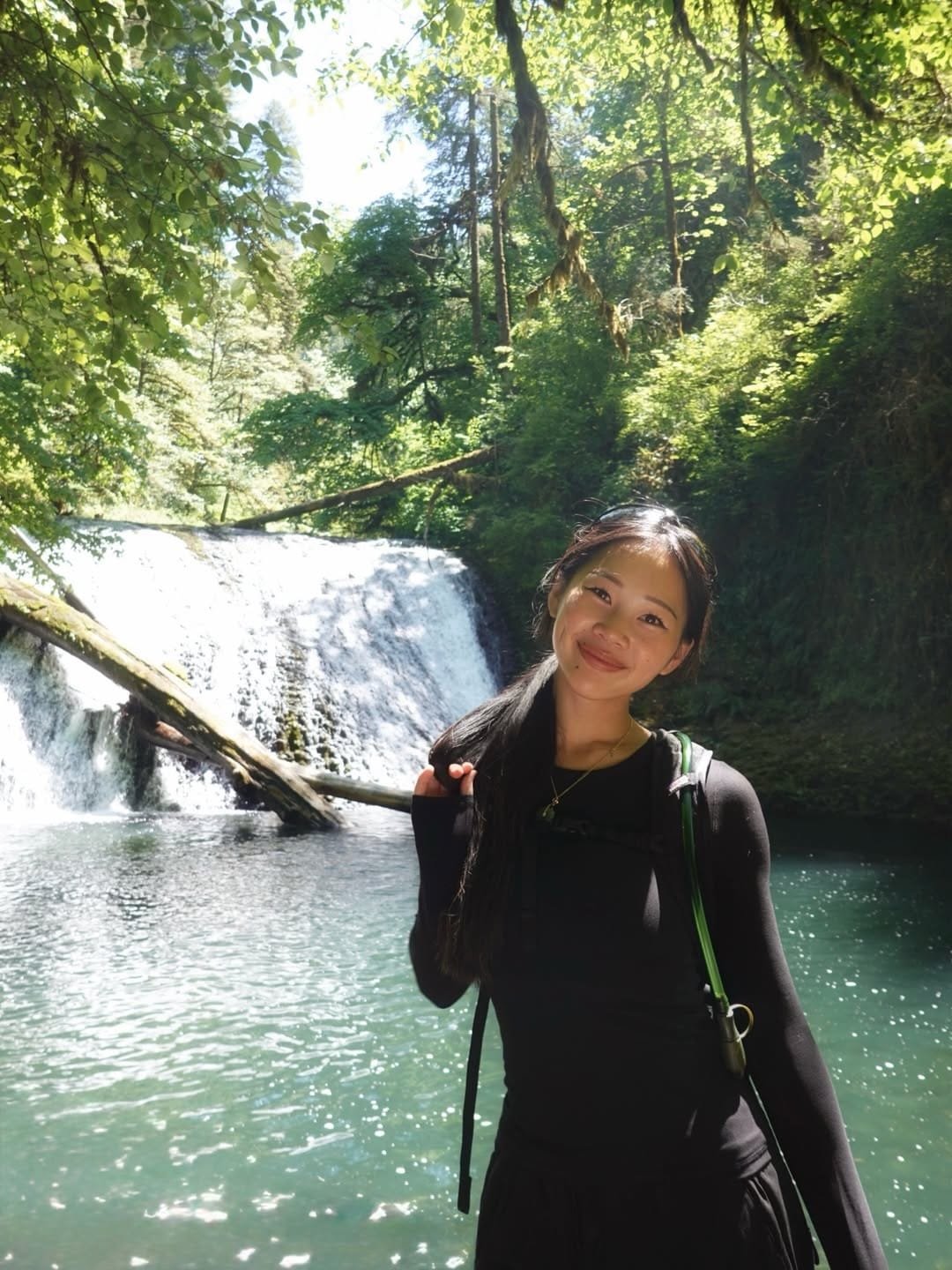 A young woman stands and smiles in front of a forest waterfall, sunlight filtering through the green trees onto the water and her face. She wears a black long-sleeve top and has a backpack strap over her shoulder.