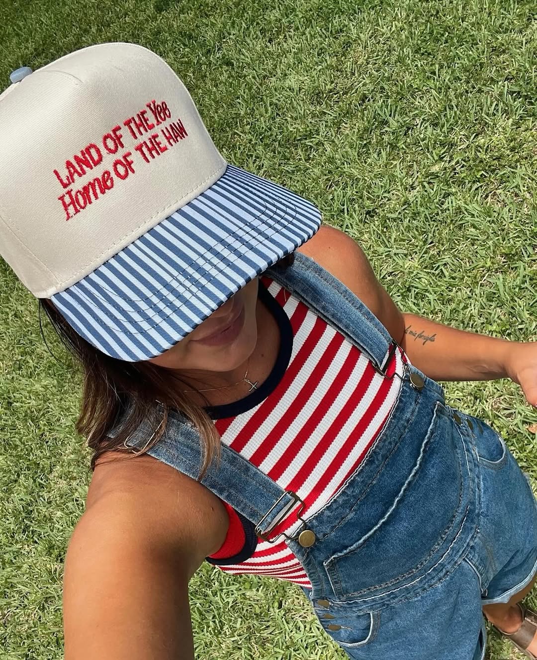 A person in a blue denim overall, red and white striped top, and a “LAND OF THE free Home OF THE Hmm” cap stands on green grass, face mostly obscured—capturing effortless Fourth of July fashion.