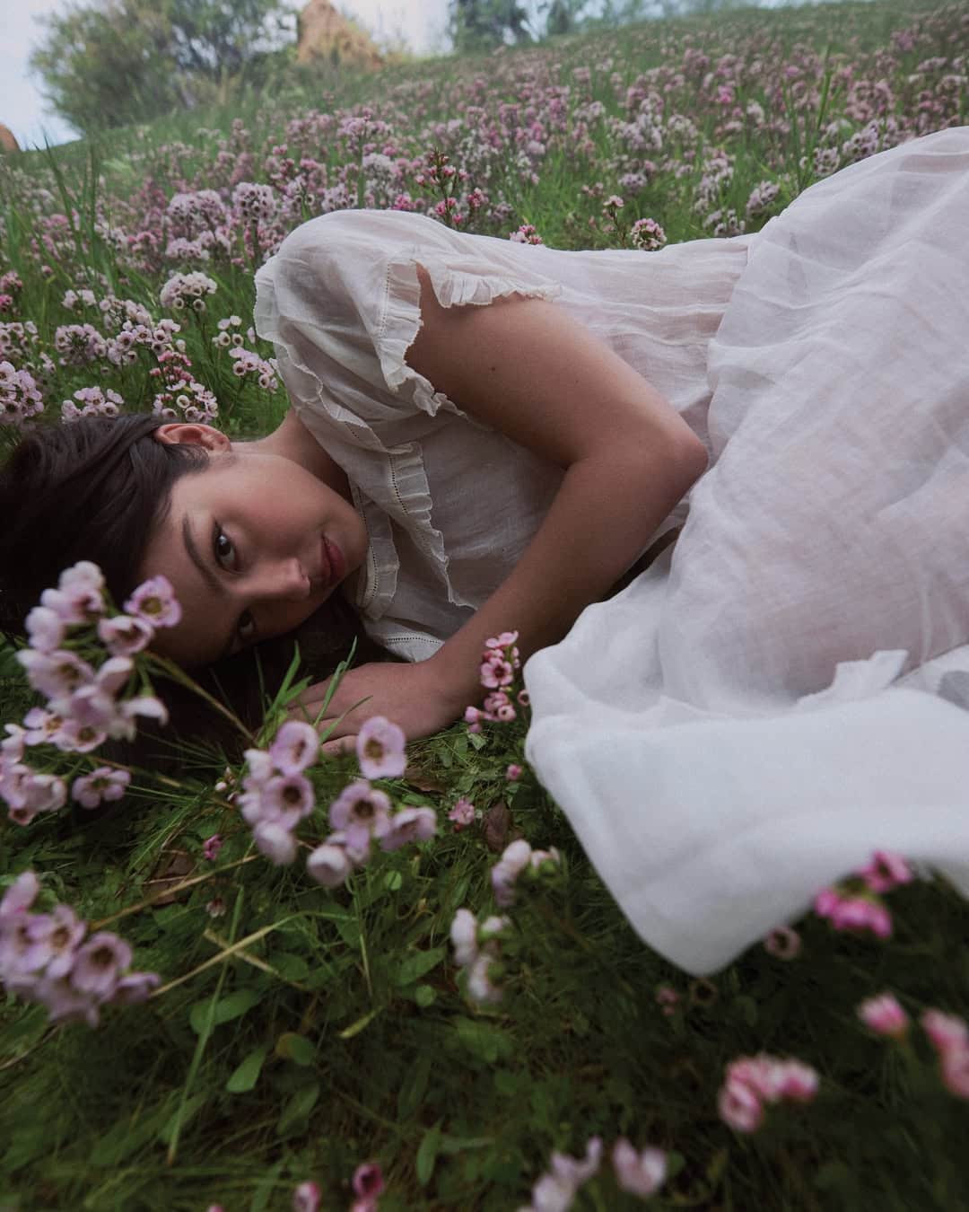 A woman in a white, sheer dress lies on her side beside a winding stream track in a field of pink wildflowers, gazing softly at the camera, surrounded by lush green grass and blooms.