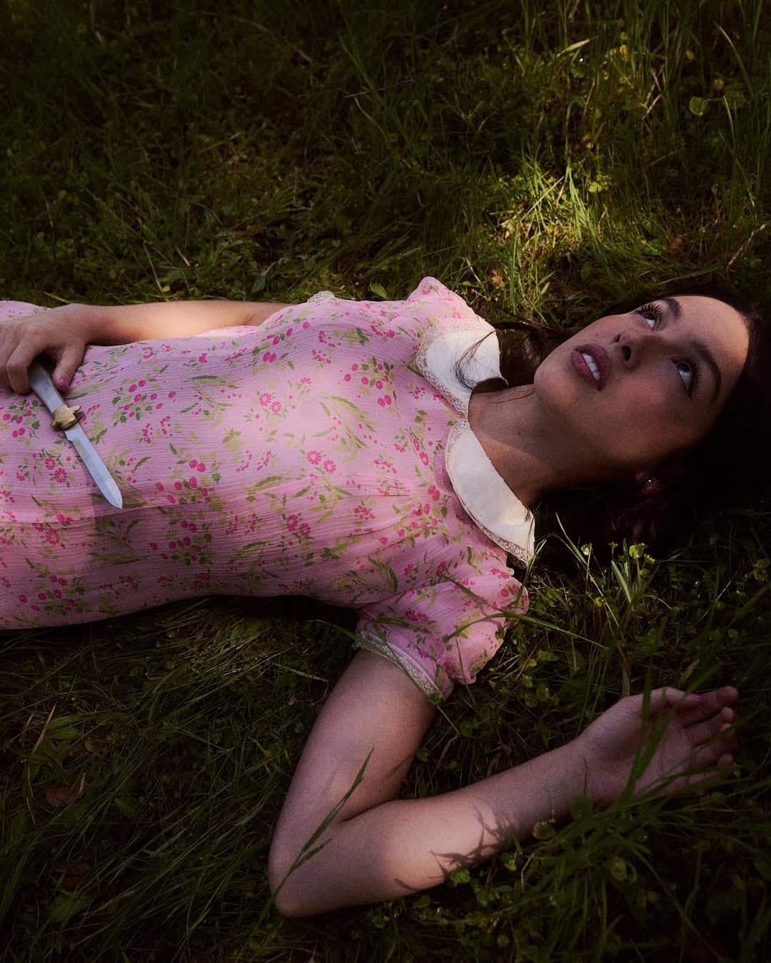 A woman in a vintage pink floral dress lies on grass by a stream, looking up with a pensive expression. She holds a small knife in her right hand, sunlight filtering through greenery—a scene from "You Seem Pretty Sad for a Girl So in Love.
