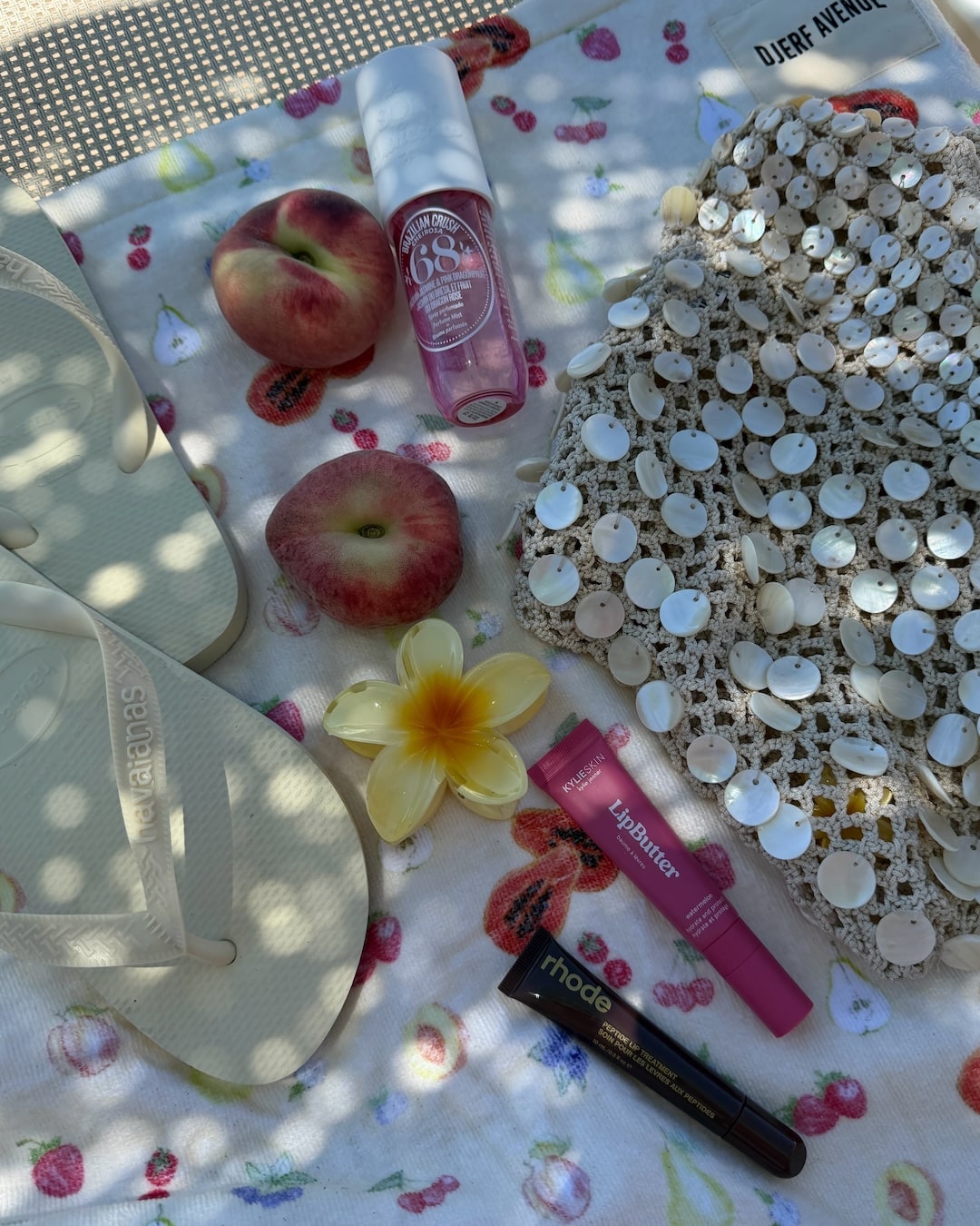 A flat lay of summer essentials: white flip-flops, a sequined bag, two flat peaches, a yellow flower, a facial mist, pink lip balm, and black mascara, all arranged on a floral-patterned towel in dappled sunlight.
