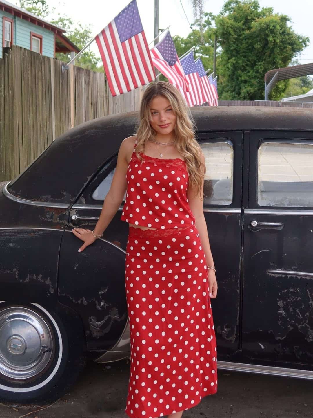A woman in a red polka dot top and skirt, a classic Fourth of July outfit, stands beside a vintage black car. Several American flags are displayed in the background near a wooden fence and trees.